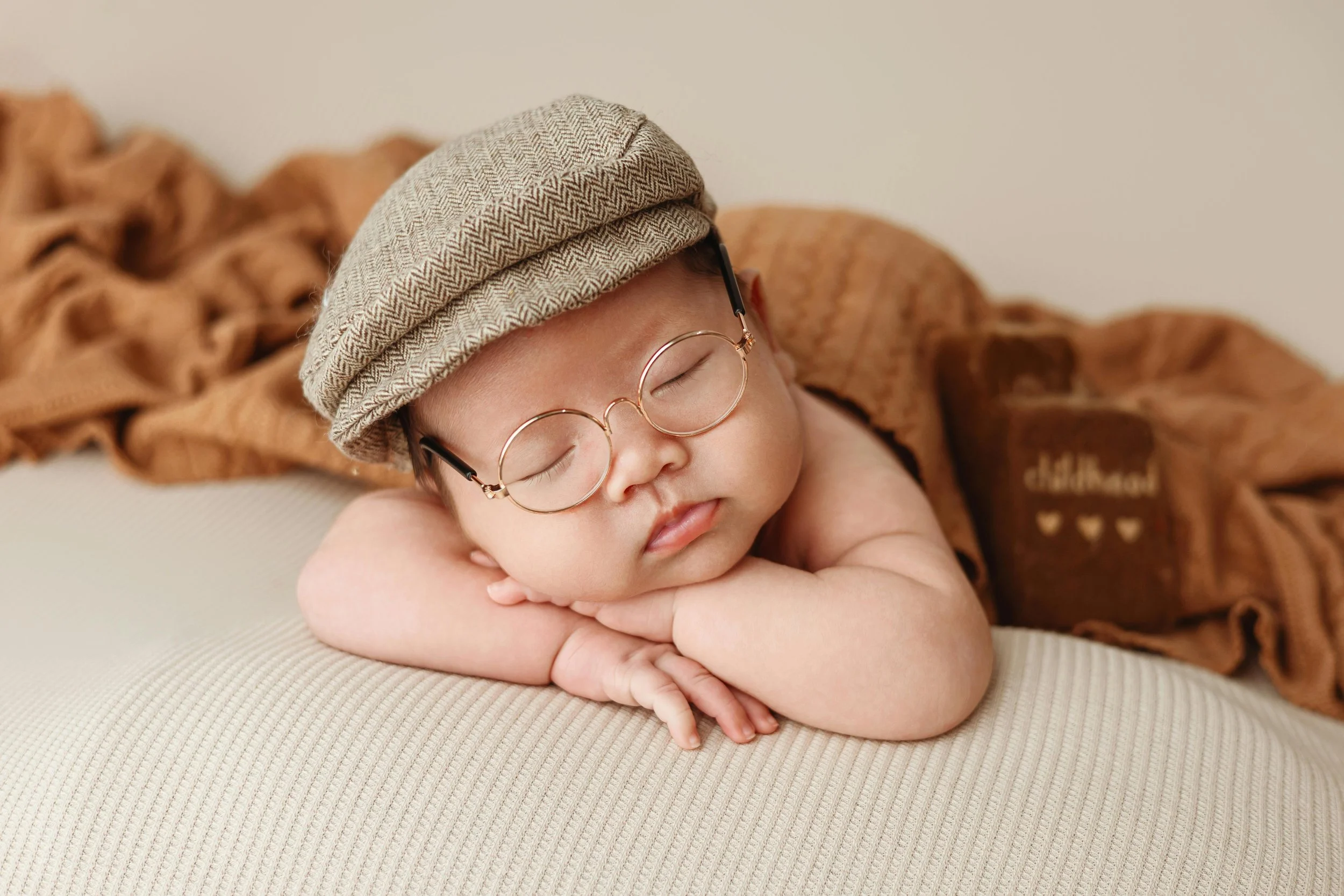 A sleeping baby with glasses and a beige cap, resting on crossed arms on a beige surface, covered partially with a brown blanket.
