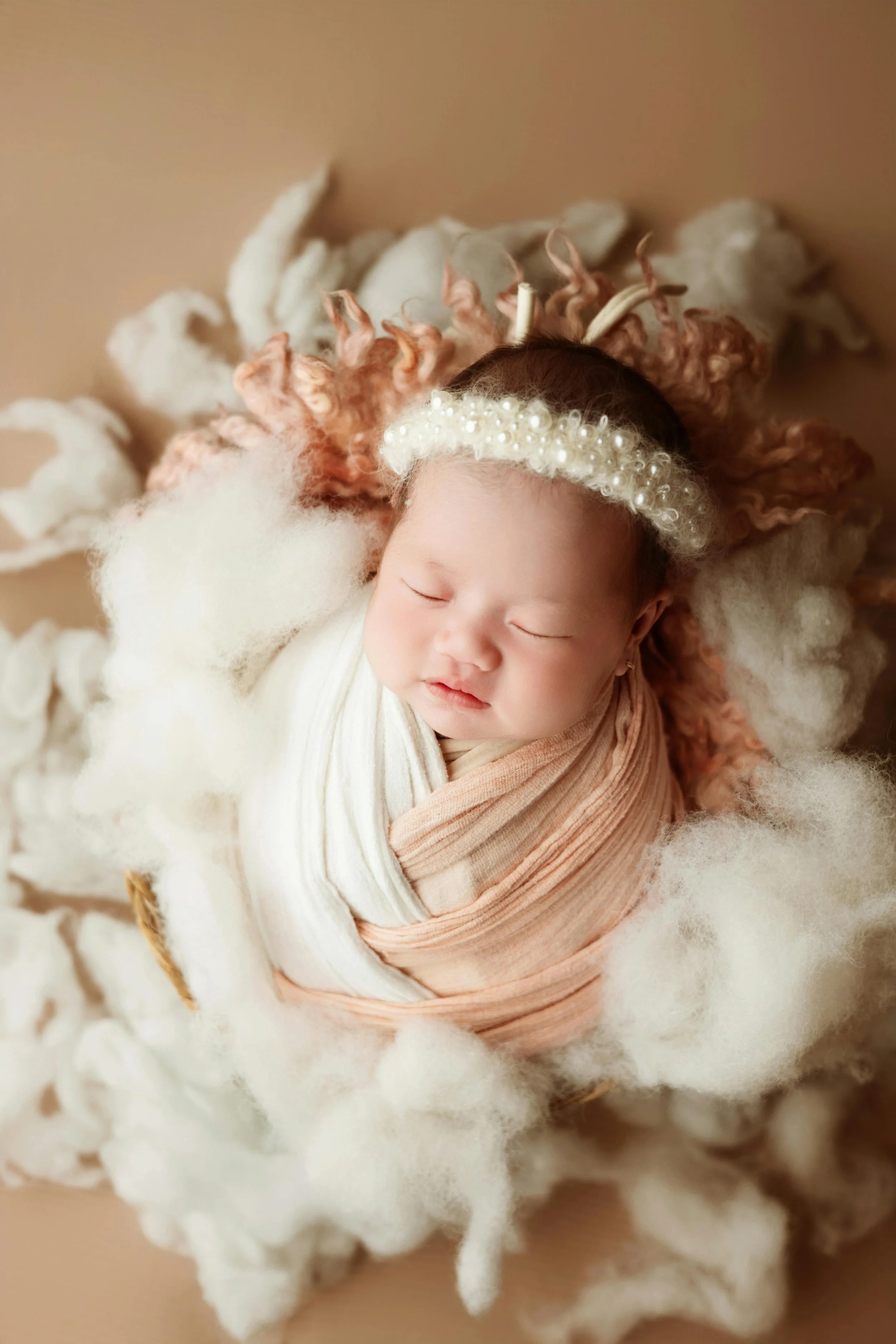 A sleeping newborn baby wrapped in a beige and white cloth, lying on a fluffy round bed with soft white and pink yarn around her head, wearing a pearl headband.