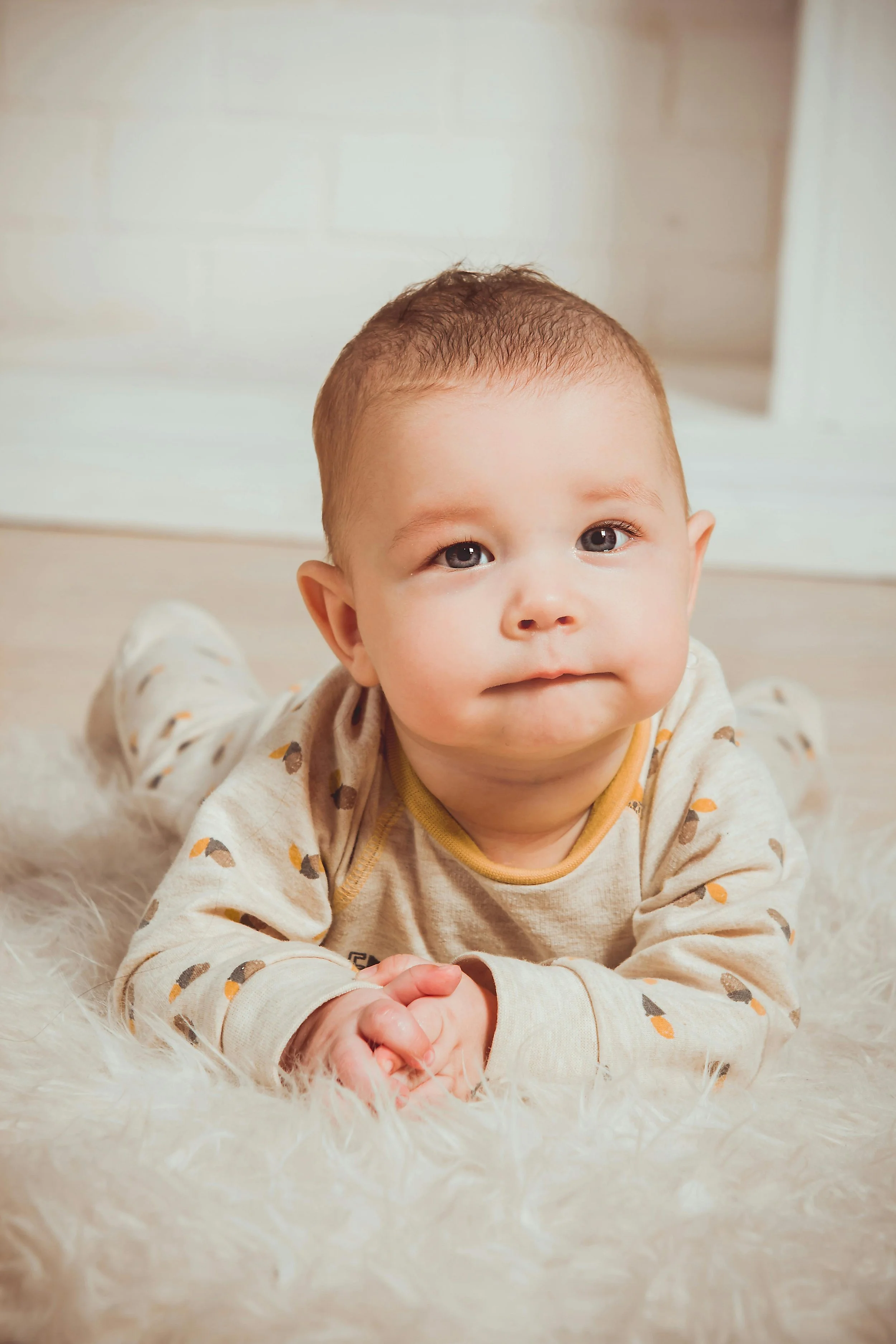 A baby with short brown hair and light skin lying on a soft, fluffy, white rug, wearing a beige onesie with a pattern of small, dark, and orange shapes, looking up at the camera with a slight smile.