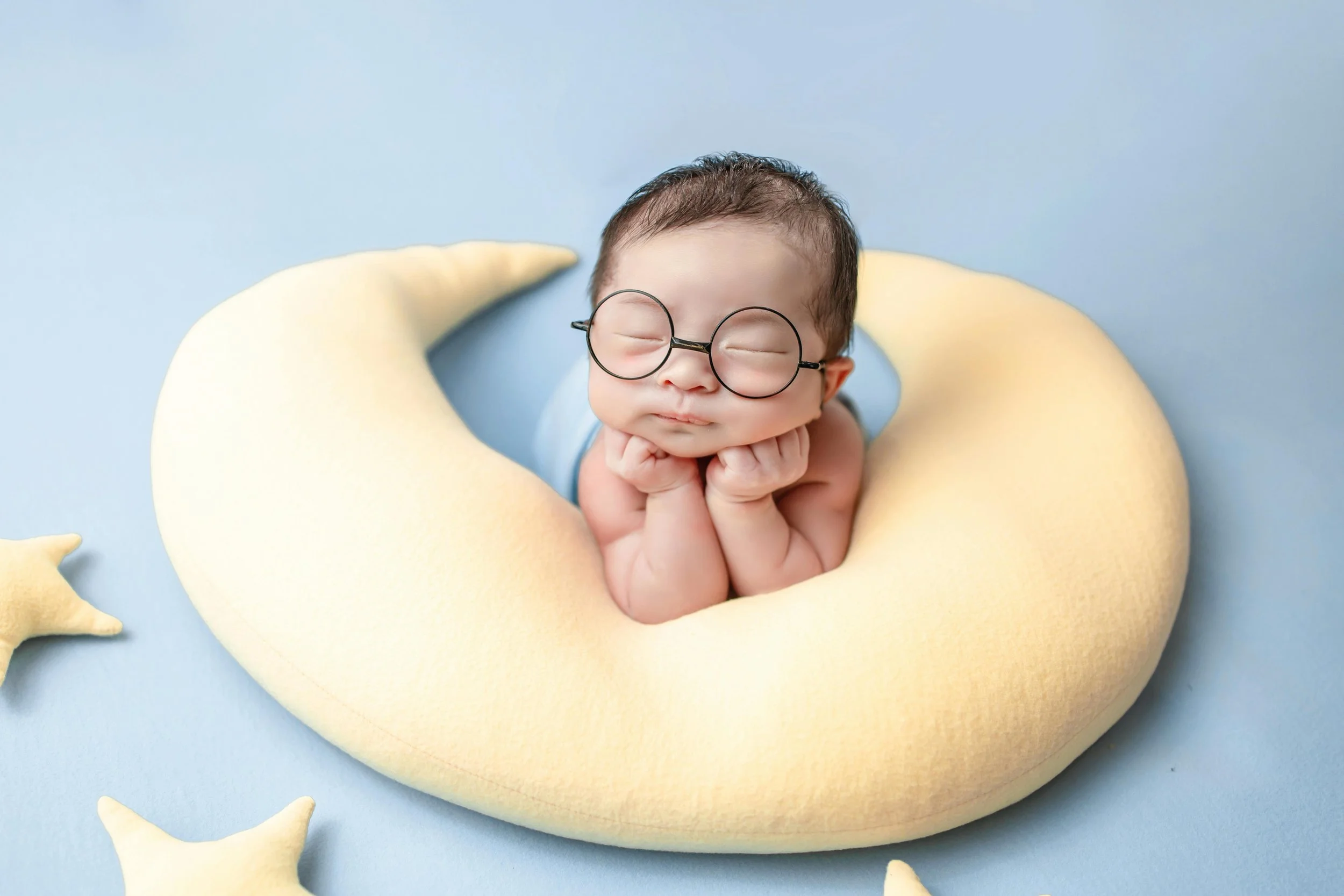 A baby with glasses resting on a yellow crescent moon-shaped pillow, surrounded by yellow stars, on a blue background.