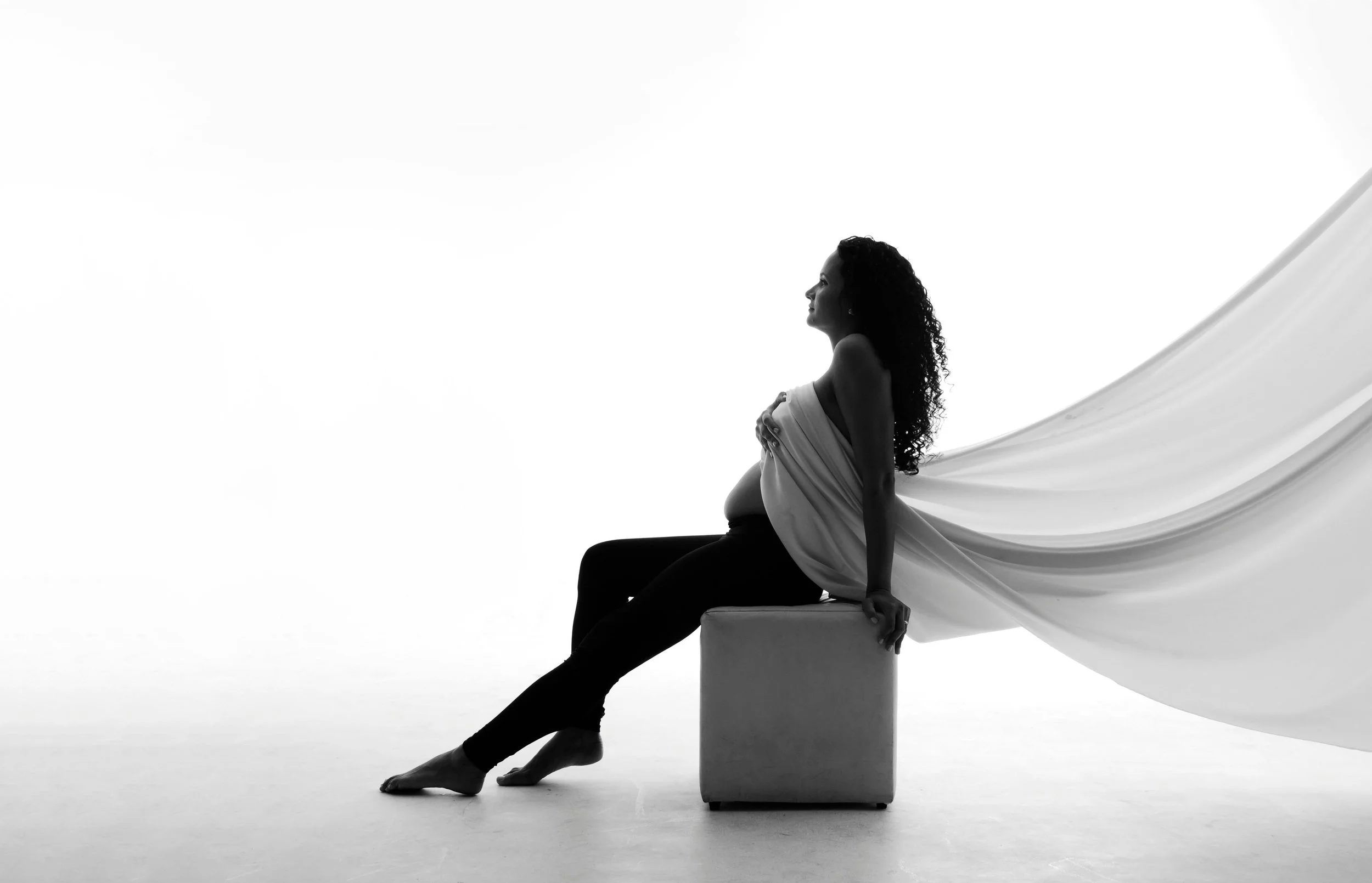 Silhouette of a pregnant woman sitting on a box with a flowing fabric cape behind her, in black and white.