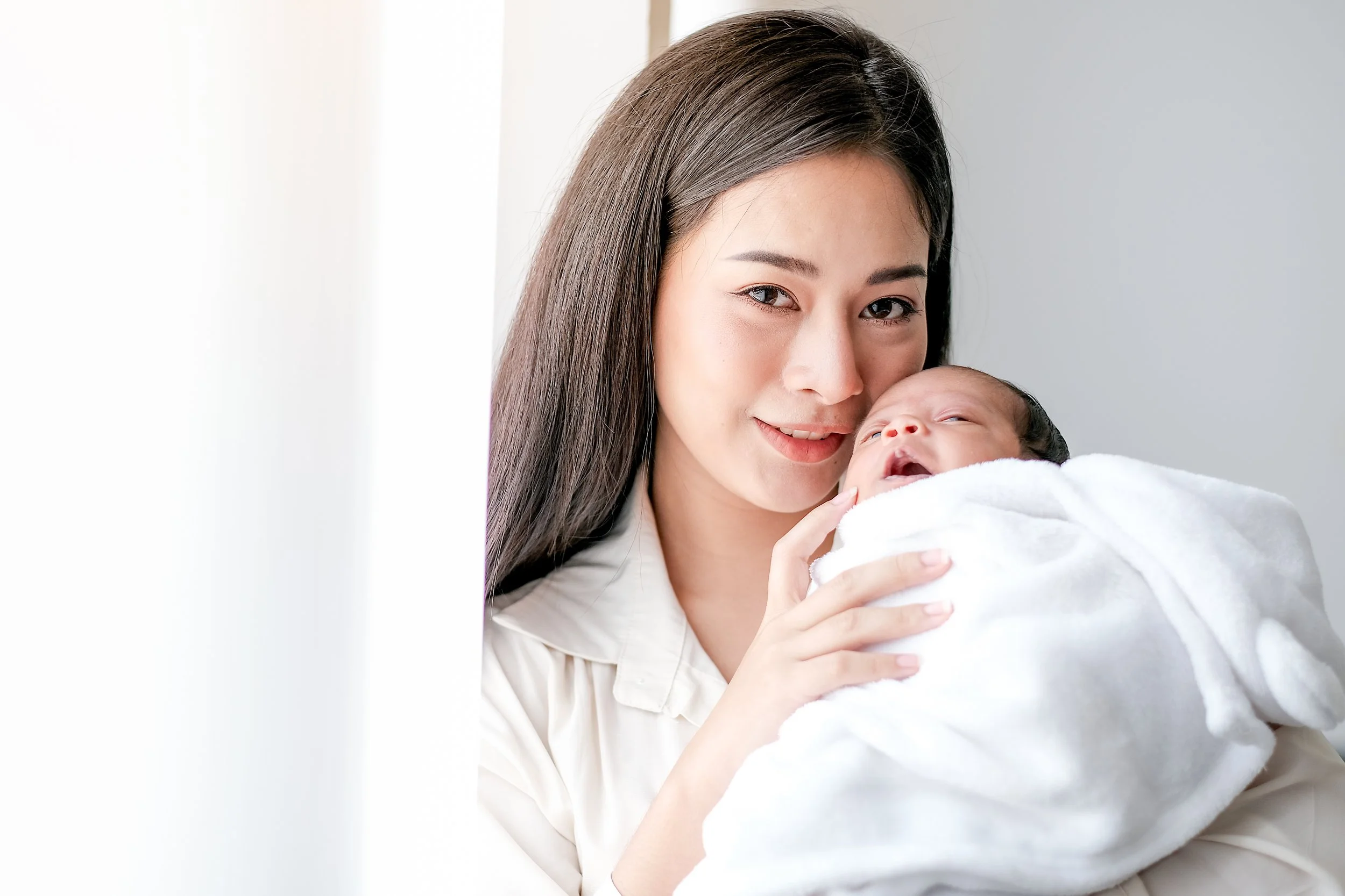 A woman holding a newborn wrapped in a white towel, both smiling softly indoors.