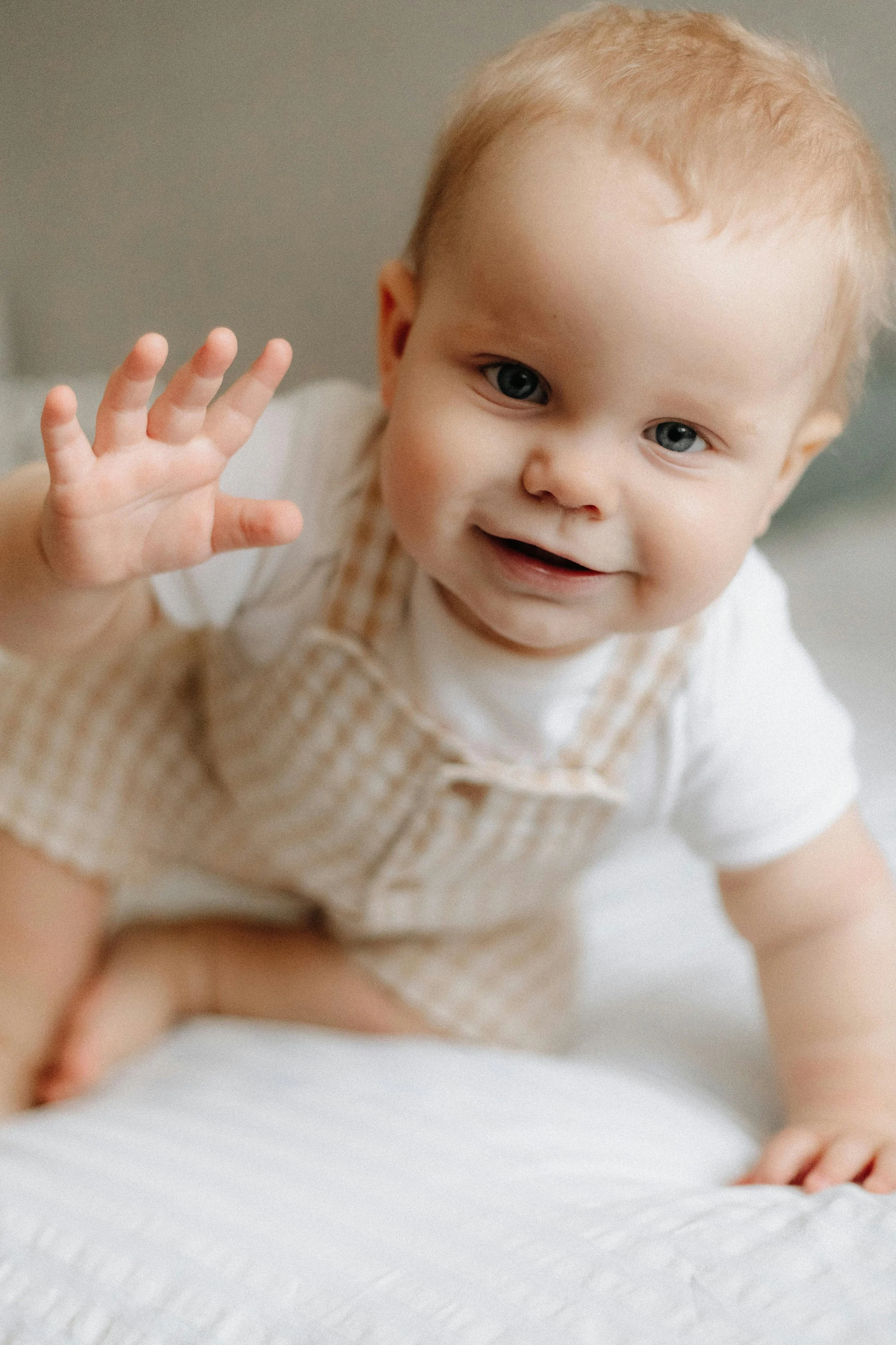 A smiling baby with blue eyes and light brown hair crawling, wearing a white shirt and checked beige overalls, waving with one hand.