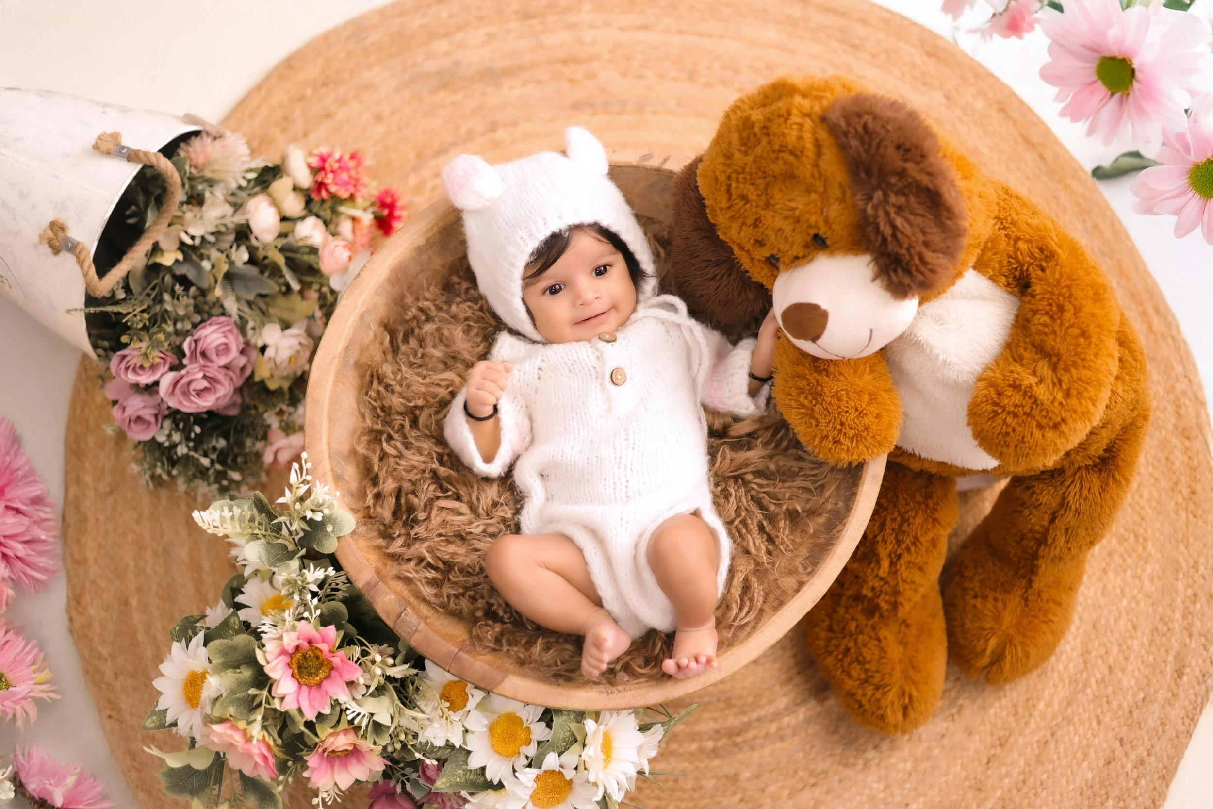 A baby in a white knitted outfit and hat with bear ears, lying in a brown furry nest with a large stuffed dog toy, surrounded by pink and white artificial flowers on a round woven rug.