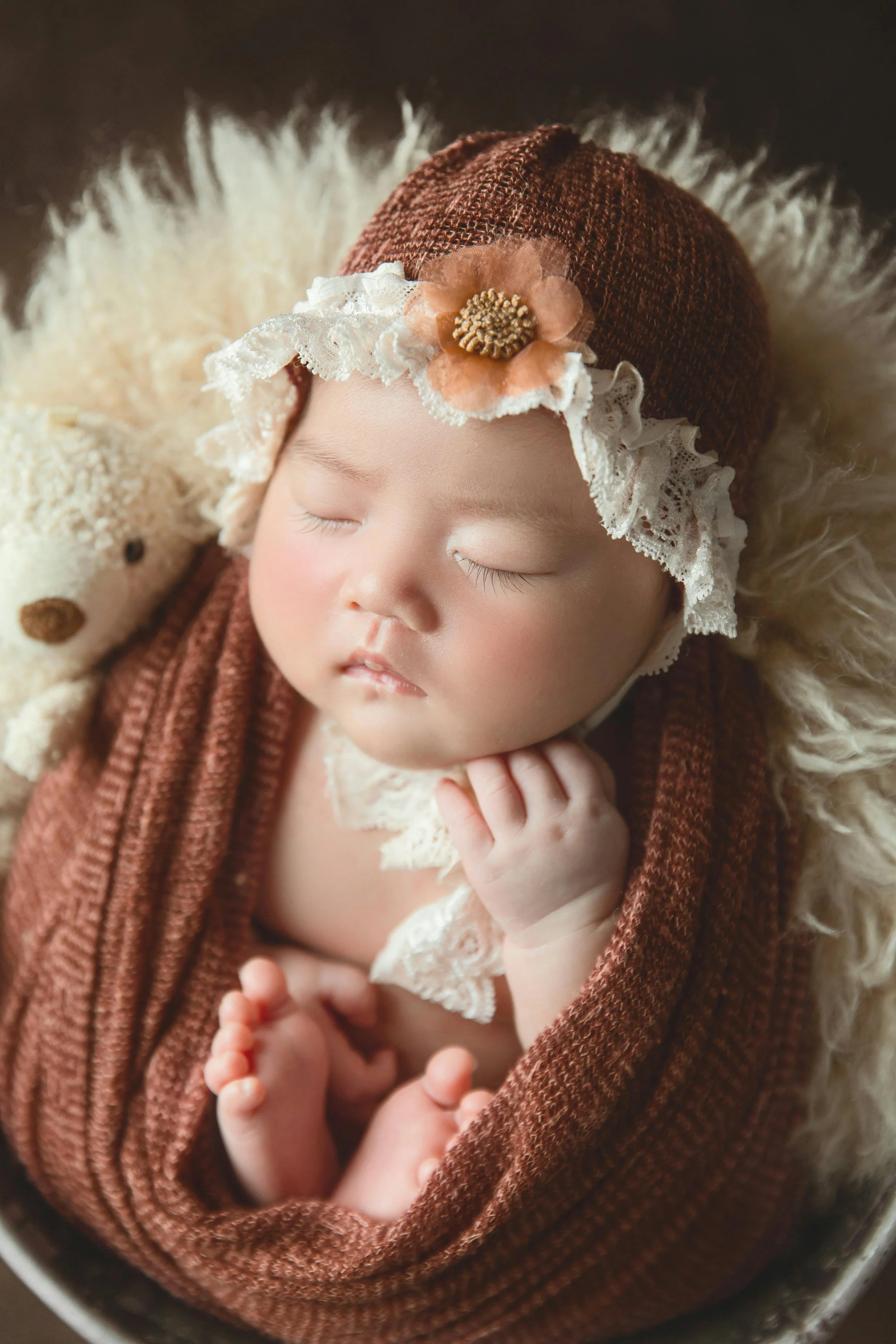 A sleeping baby wrapped in a brown knit blanket, wearing a lace headband with an orange flower, nestled on a fluffy beige surface, with a cream-colored teddy bear nearby.