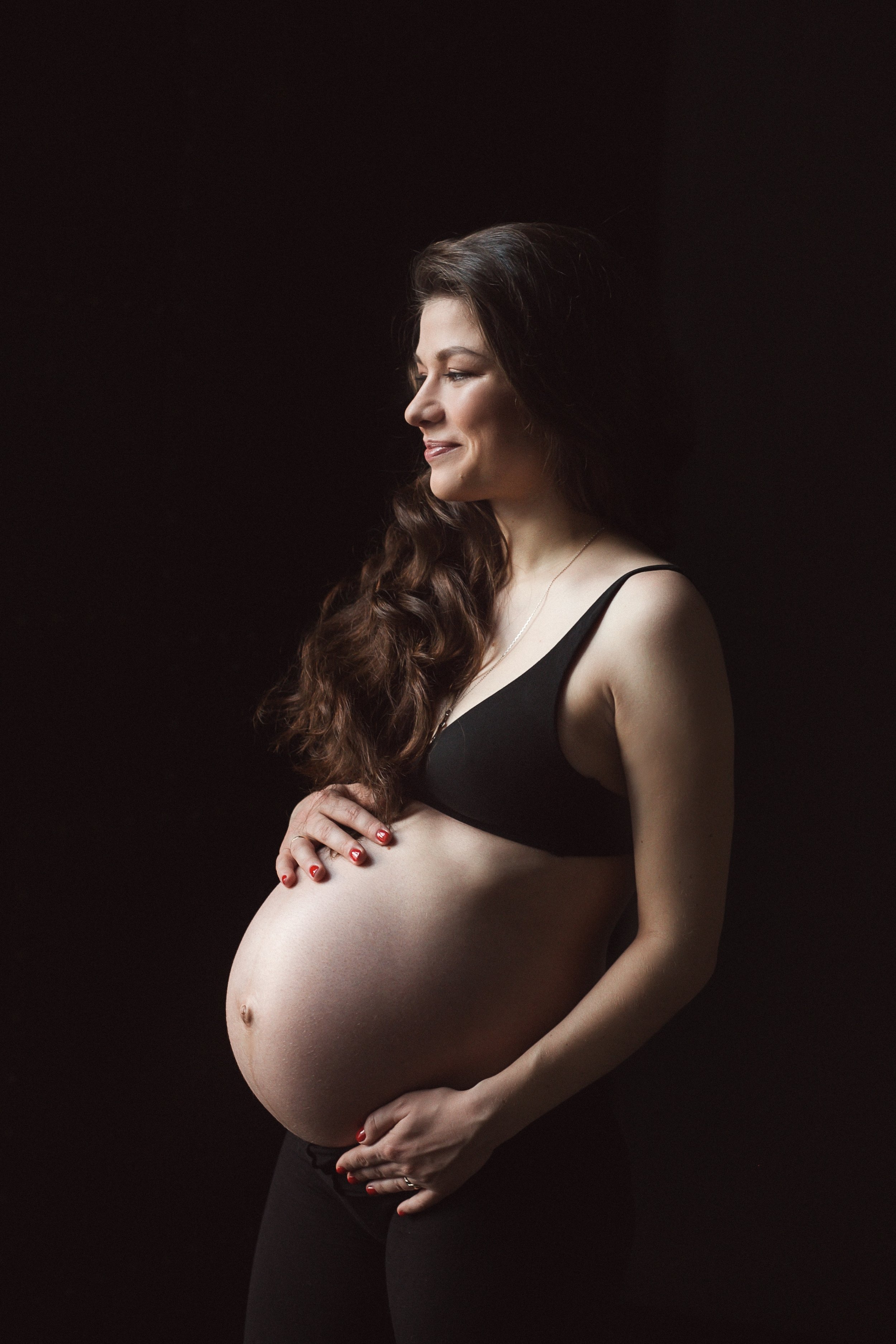 A pregnant woman with long, wavy hair wearing a black bra and pants, standing against a dark background, smiling softly and holding her belly.
