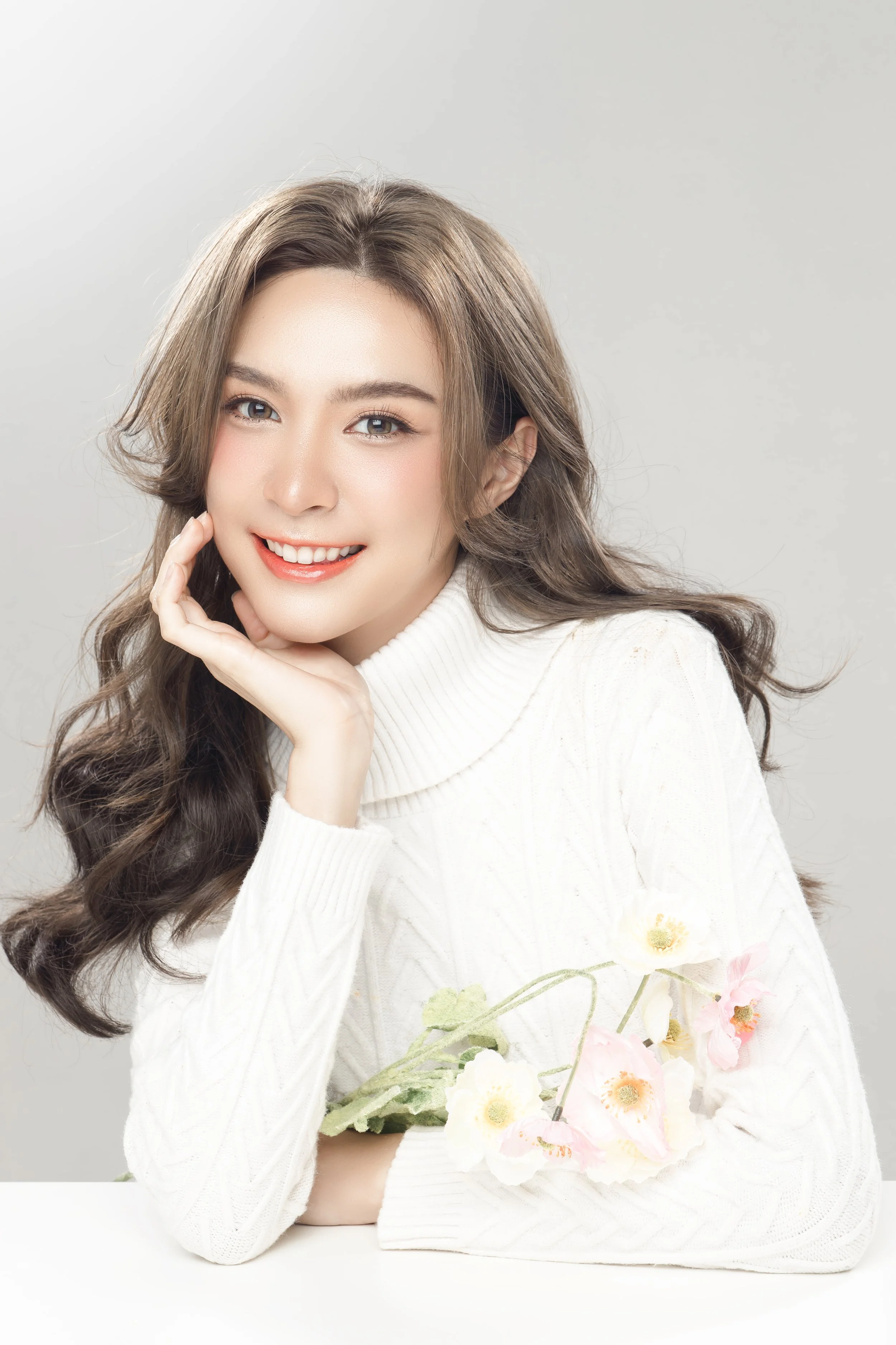 A young woman with light brown hair wearing a cream-colored sweater, smiling and resting her chin on her hand, holding pink and white flowers in her arm, against a gray background.