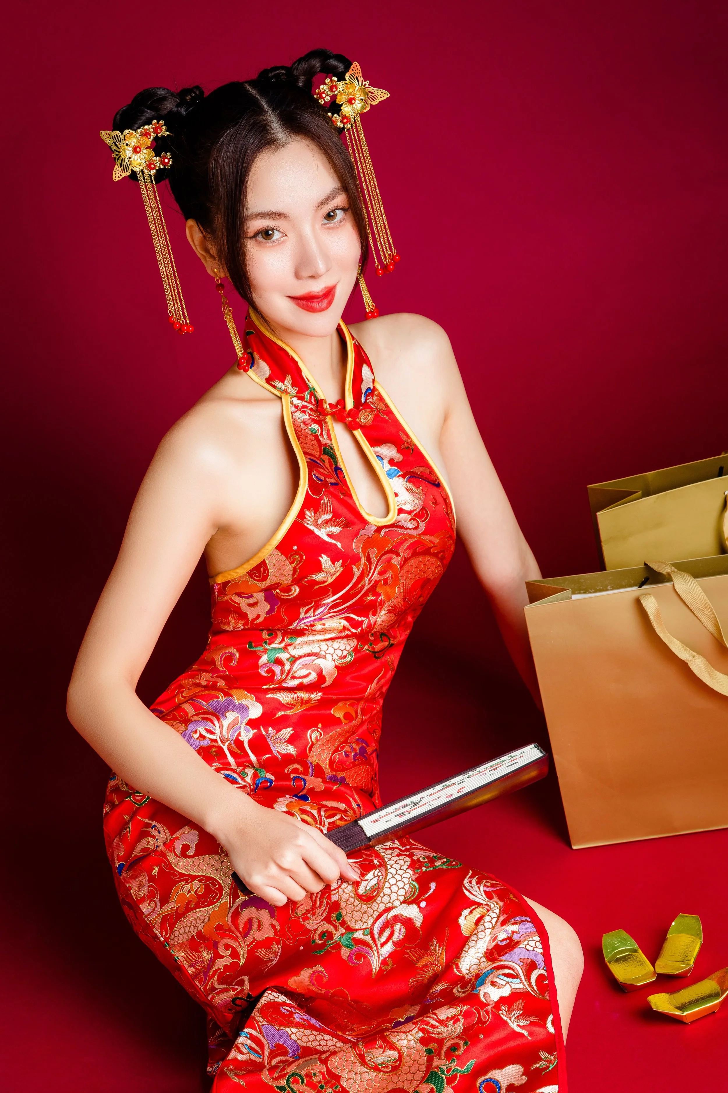 A woman dressed in a traditional red Chinese dress sitting against a red background, holding a fan, with gold shopping bags nearby, and gold shoes on the floor.