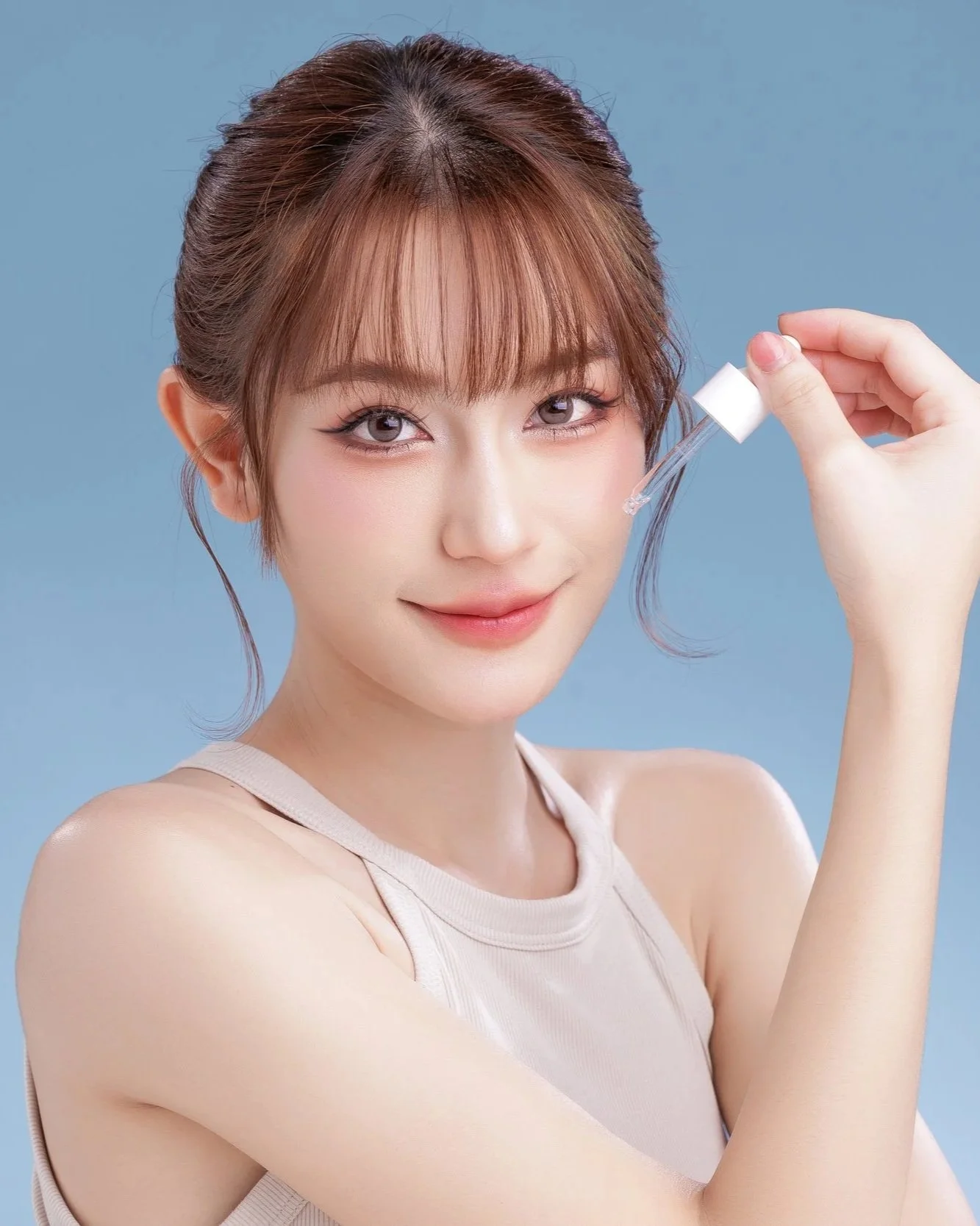 A young woman with light skin, brown hair, and bangs, holding a dropper, smiling at the camera against a plain blue background.