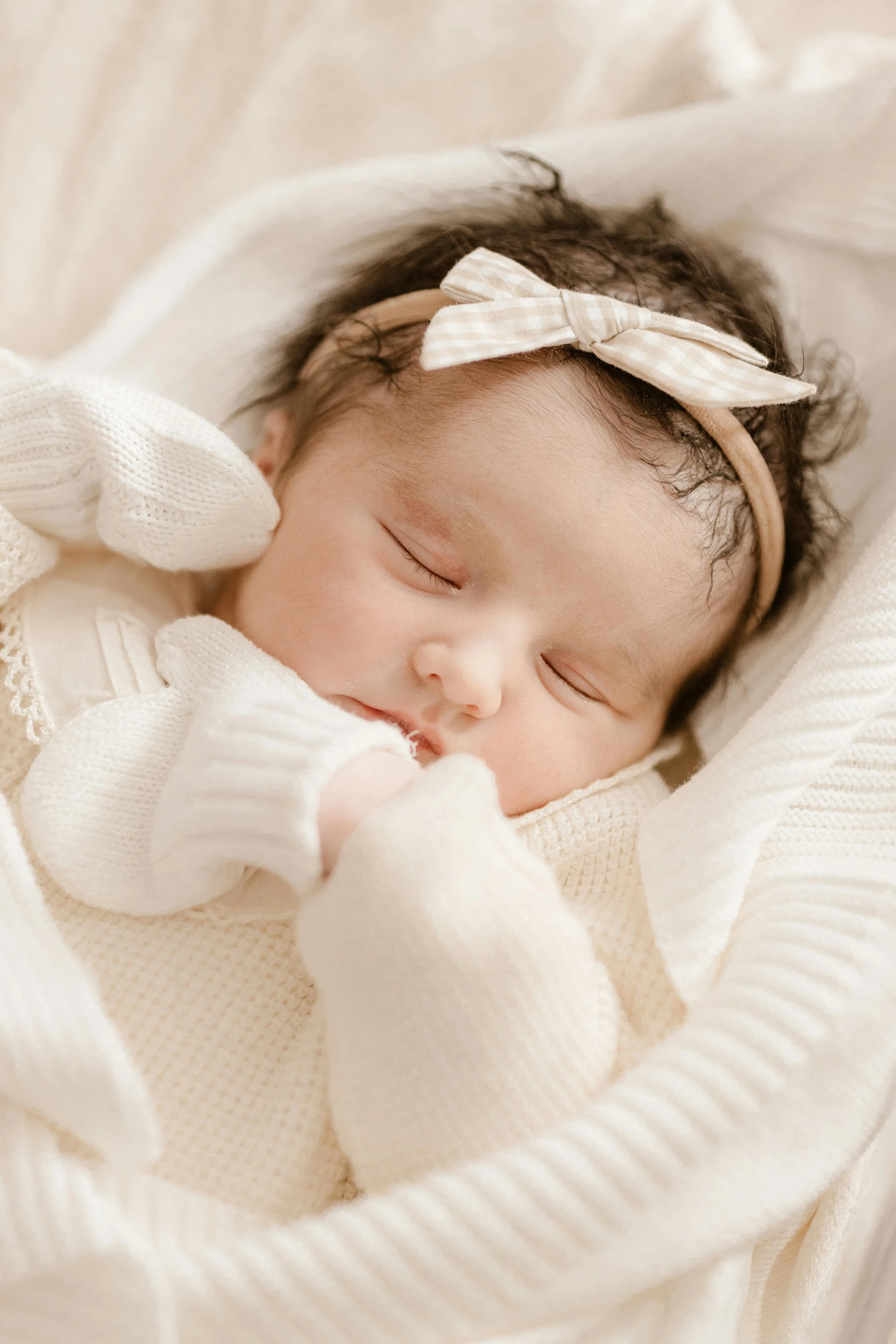 A sleeping baby with a beige headband and bow, wearing cream-colored knitted clothes, lying on a soft, white blanket.