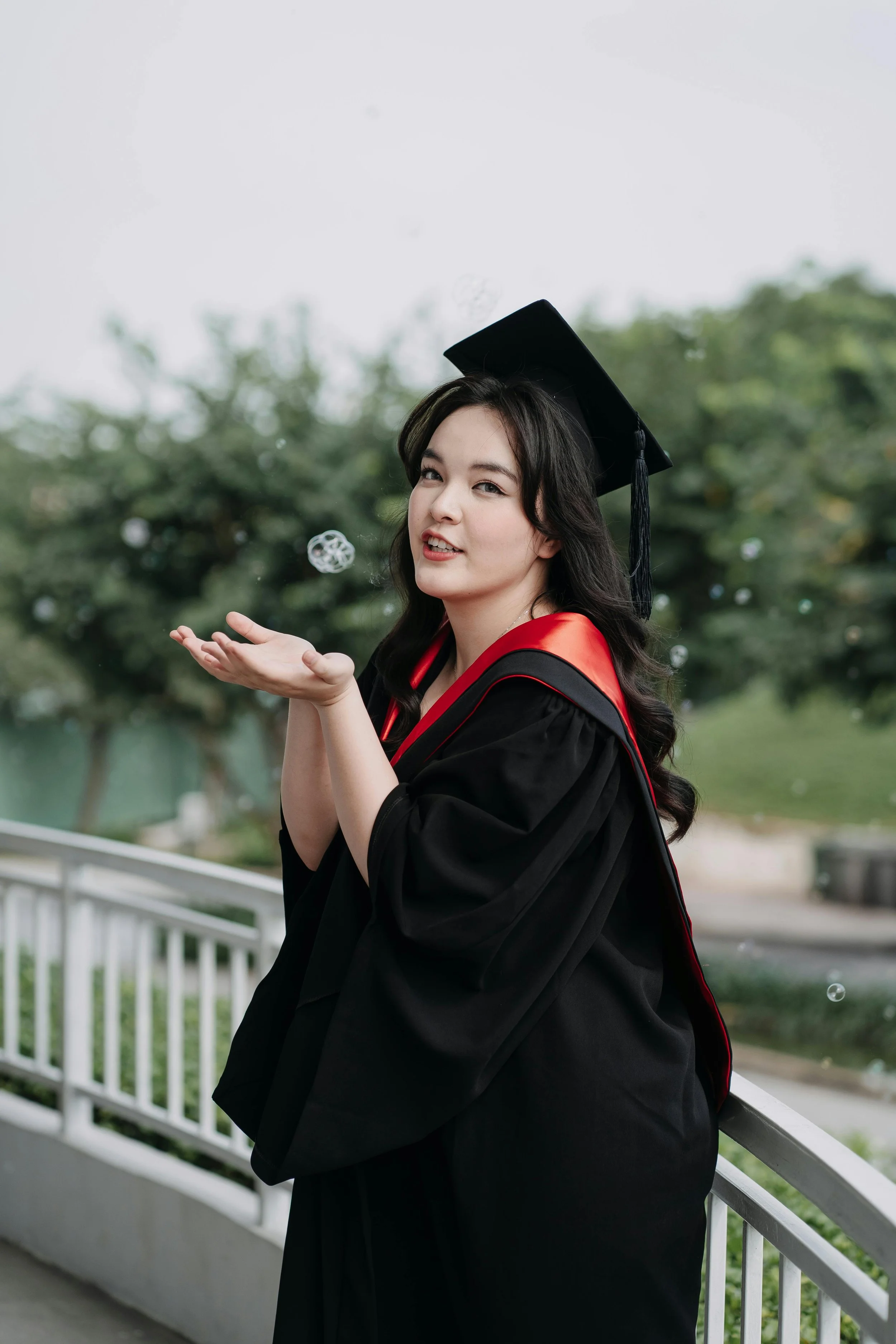 A woman in a black graduation gown and cap with a tassel, standing outdoors near a white railing. She is wearing a red stole and playing with floating bubbles while smiling and winking at the camera. Trees and a cloudy sky are in the background.