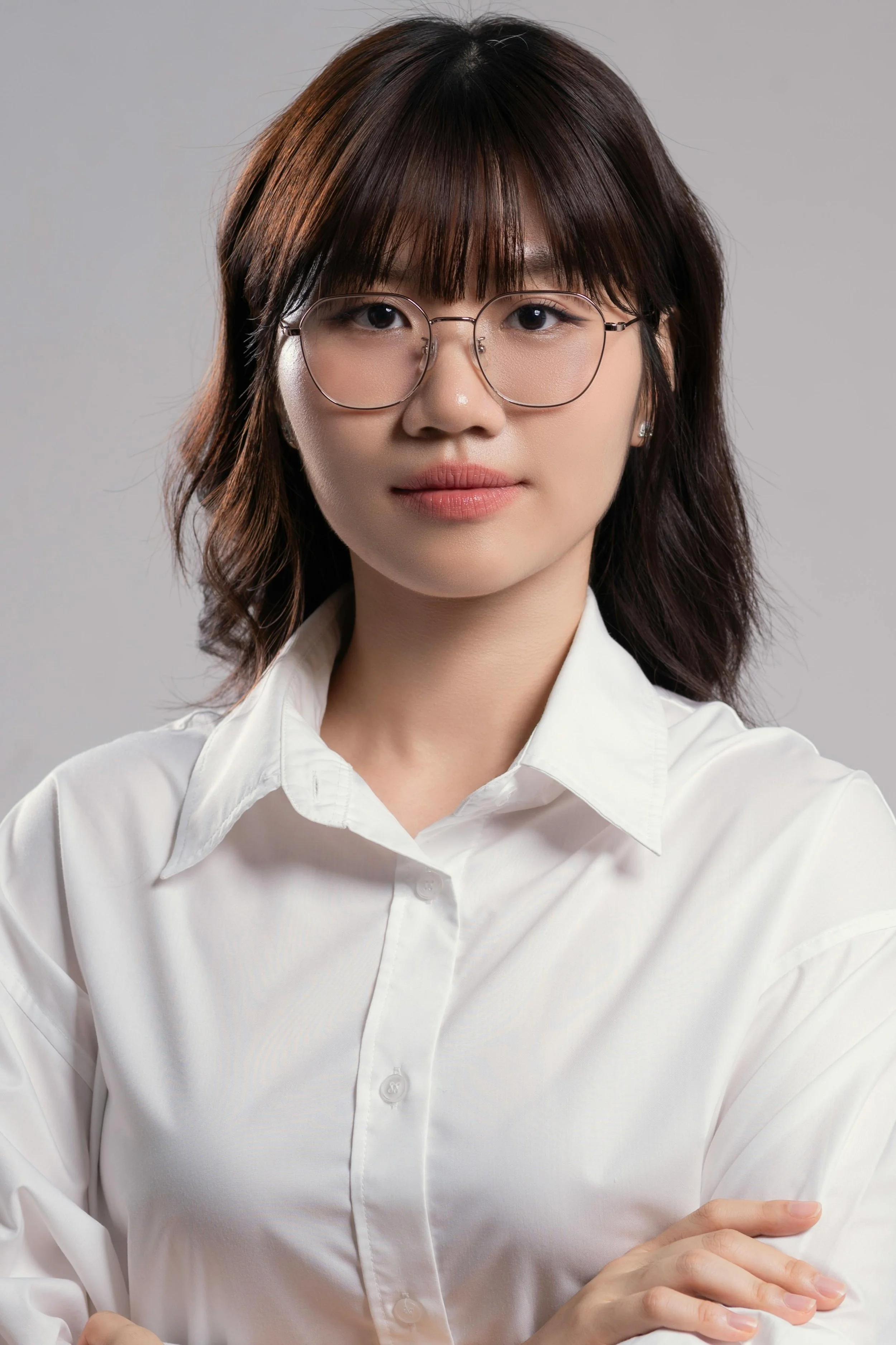 A young woman with glasses, shoulder-length brown hair, wearing a white collared shirt, standing against a light gray background.