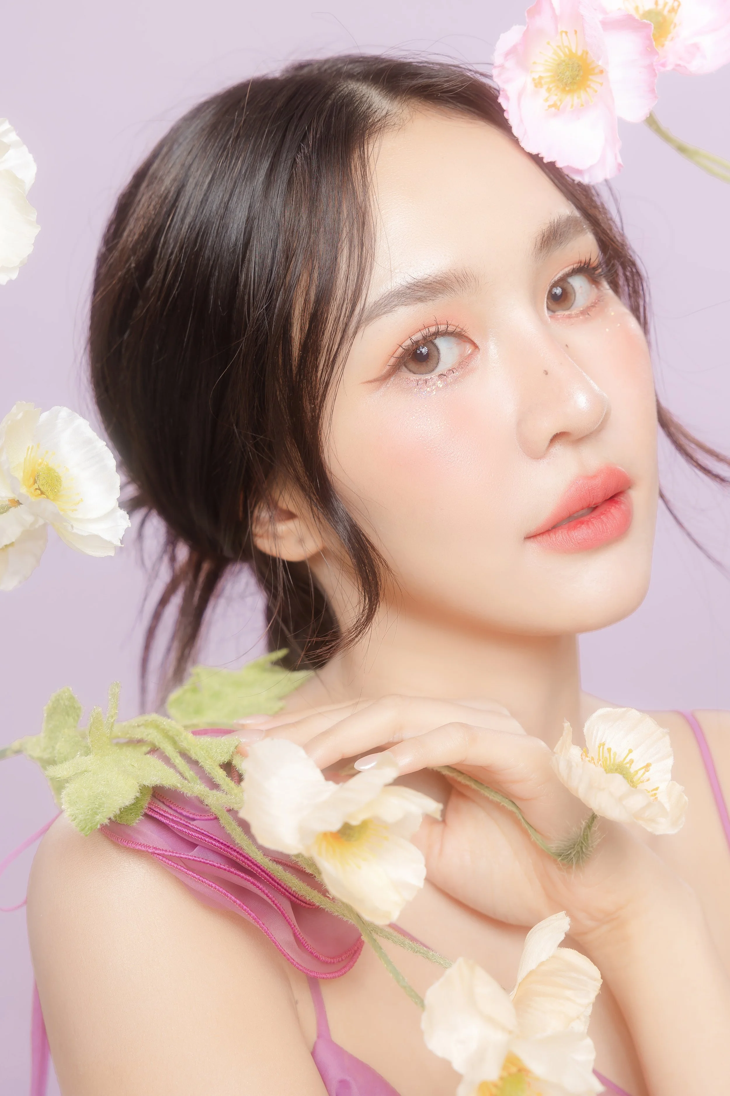 A young woman with short dark hair, wearing a pink top, surrounded by white and pink flowers with a light purple background.