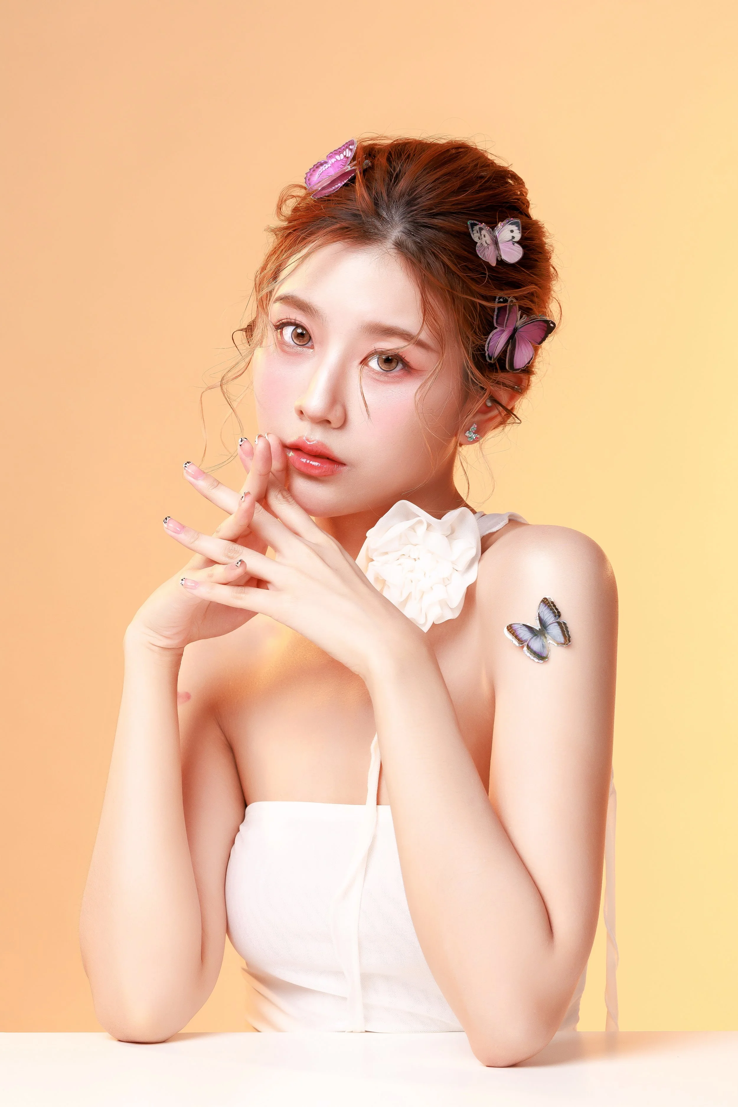 A woman with wavy hair decorated with purple and pink butterflies, wearing earrings and a white strapless top, sitting against a yellow background.