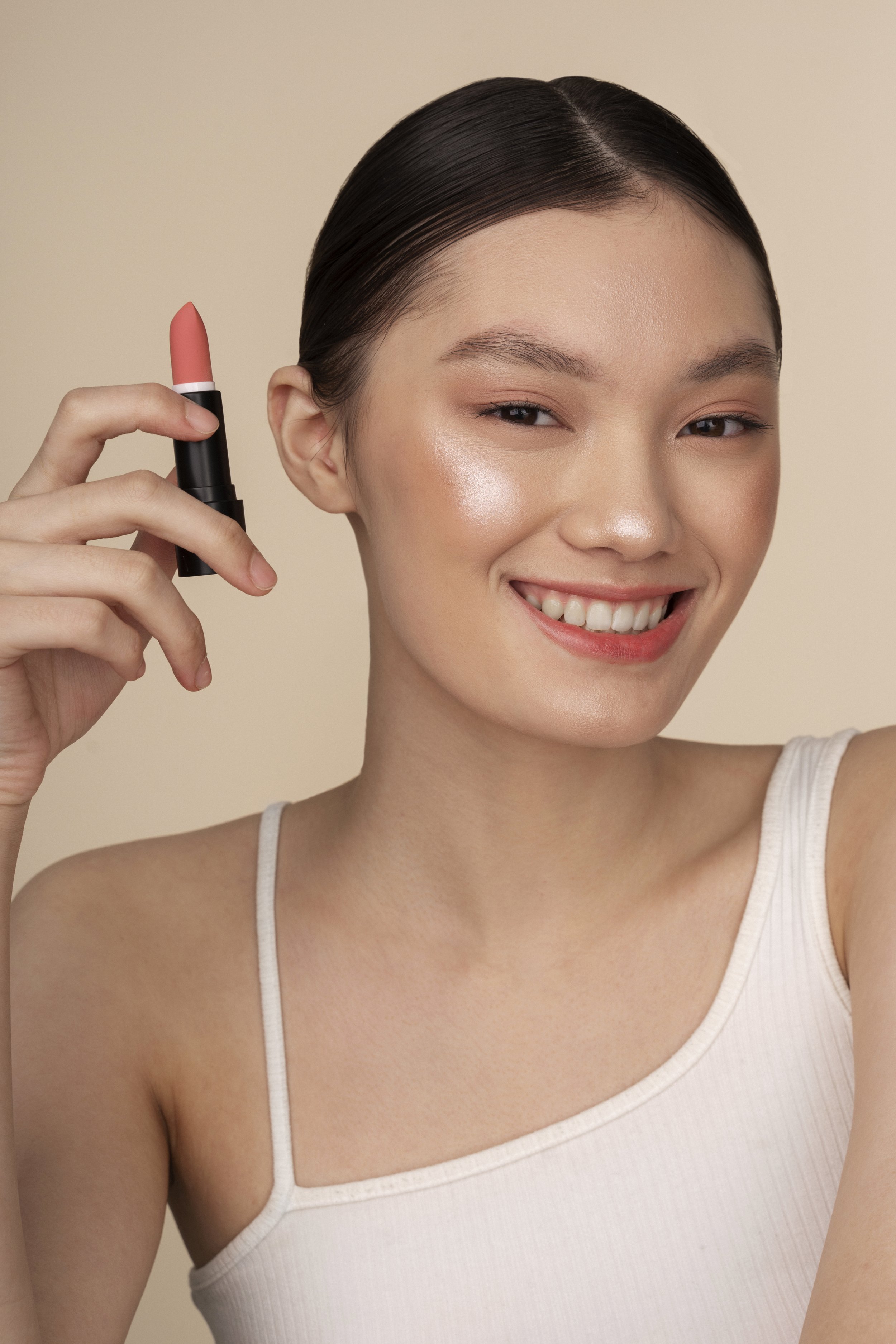 A woman smiling and holding a tube of pink lipstick near her face.
