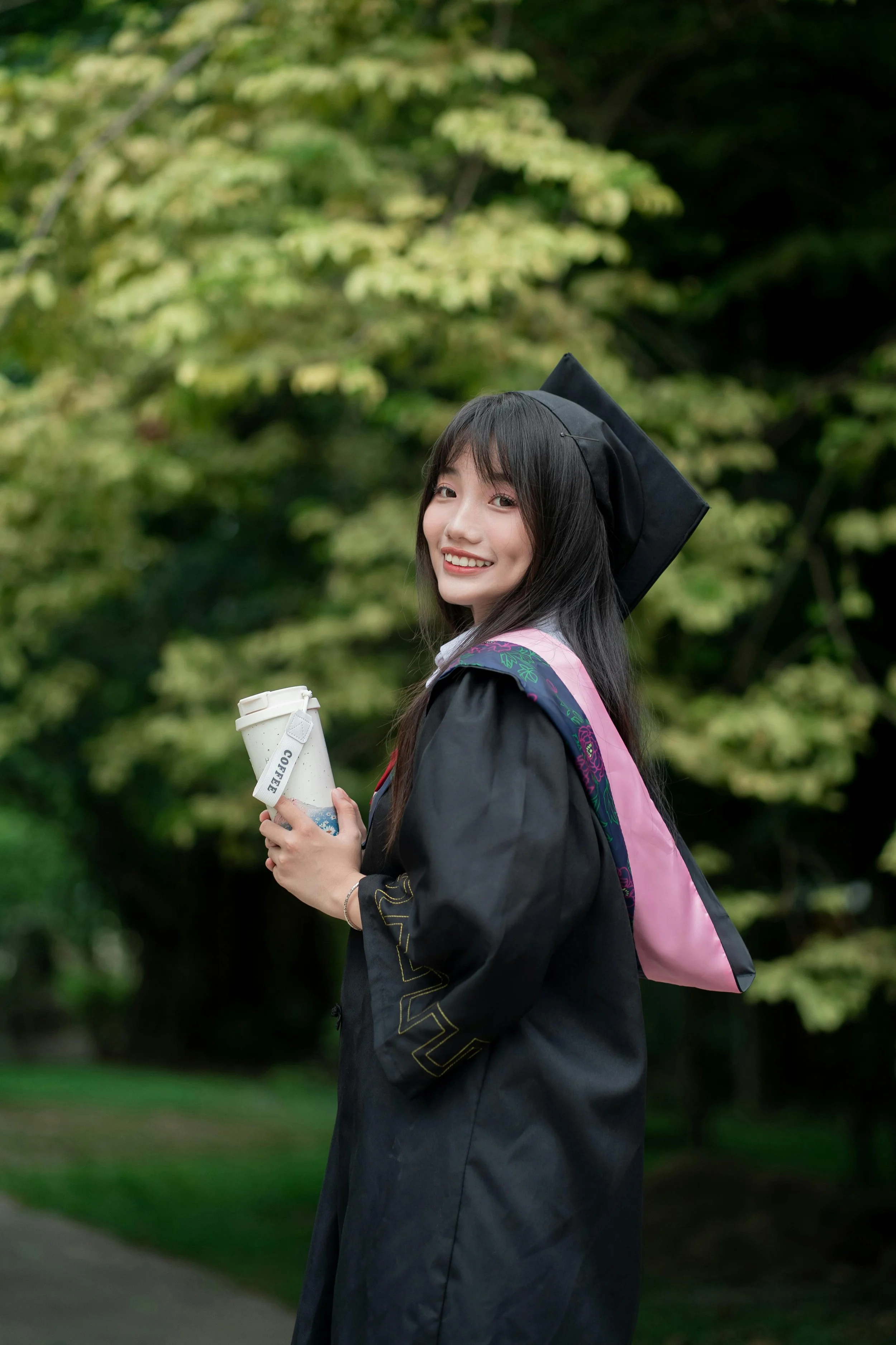 Young woman in a graduation gown and cap holding a coffee cup, smiling outdoors with trees in the background.