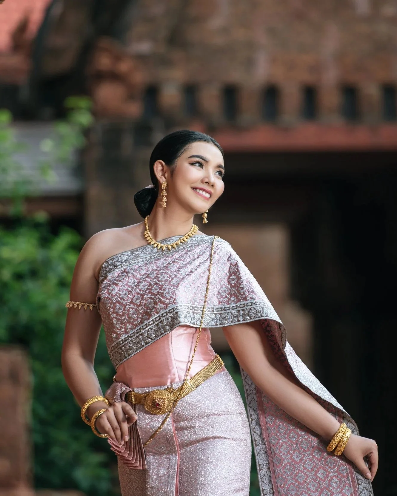 A smiling woman wearing traditional Thai attire, including a pink and silver silk dress with intricate patterns, gold jewelry, and a sash, standing outdoors.