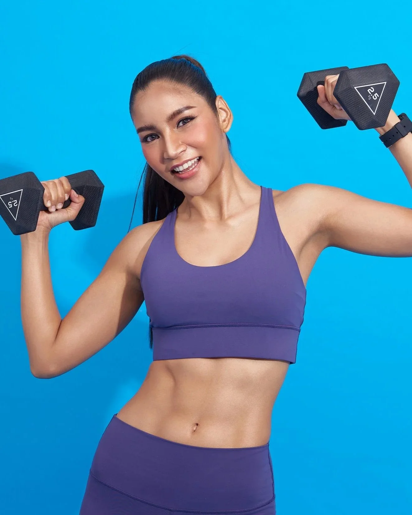 Young woman in purple sports outfit lifting black dumbbells with a bright blue background.