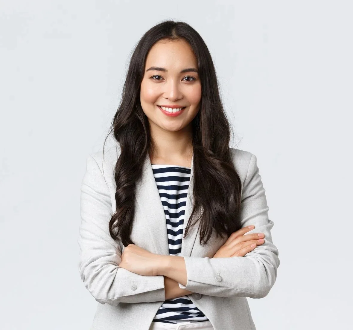 Young woman with long, dark wavy hair wearing a light blazer over a striped shirt, smiling with arms crossed against a plain light background.