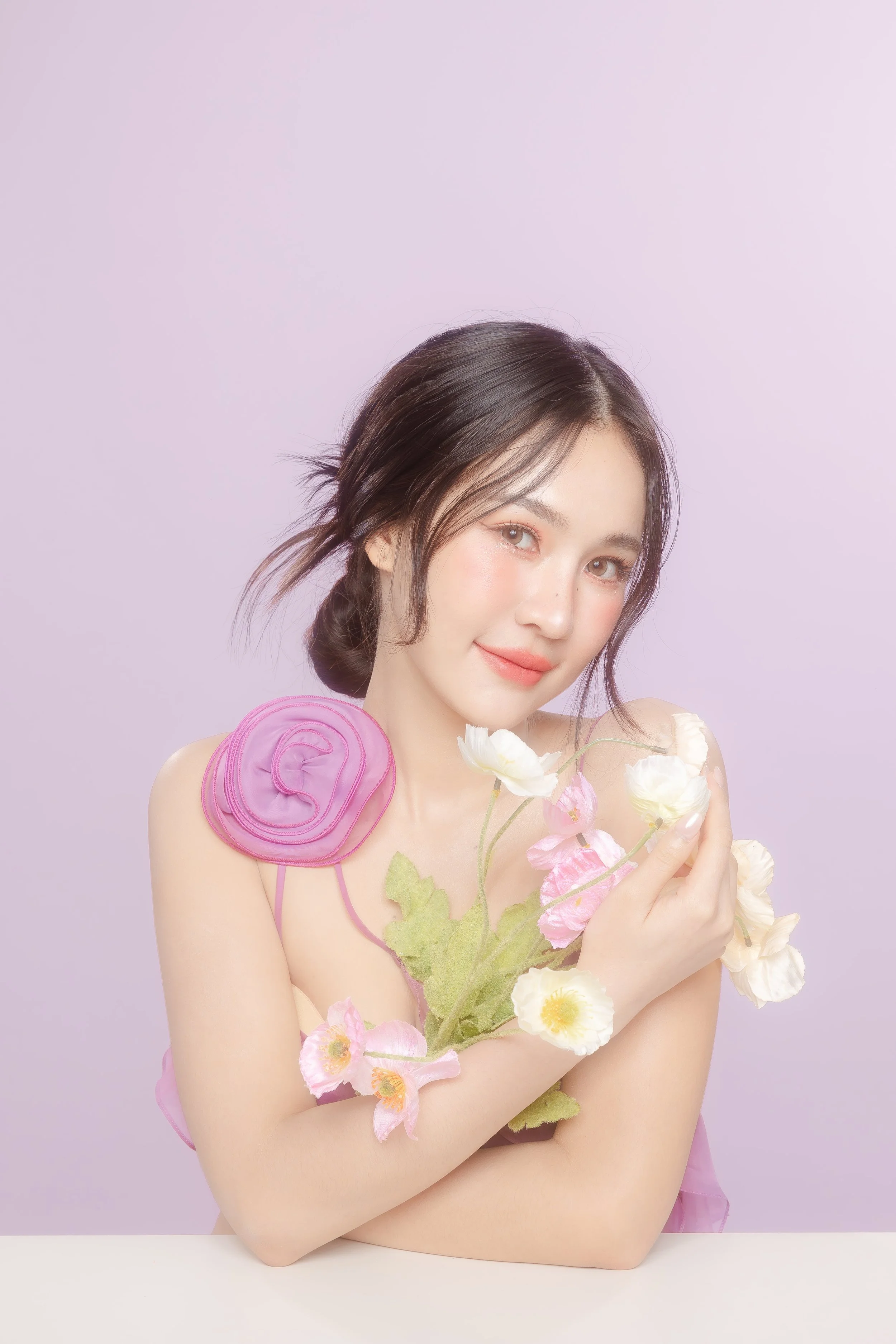 A young woman with dark hair in a loose bun, holding pastel-colored flowers, wearing a pink ruffled shoulder accessory, against a light purple background.
