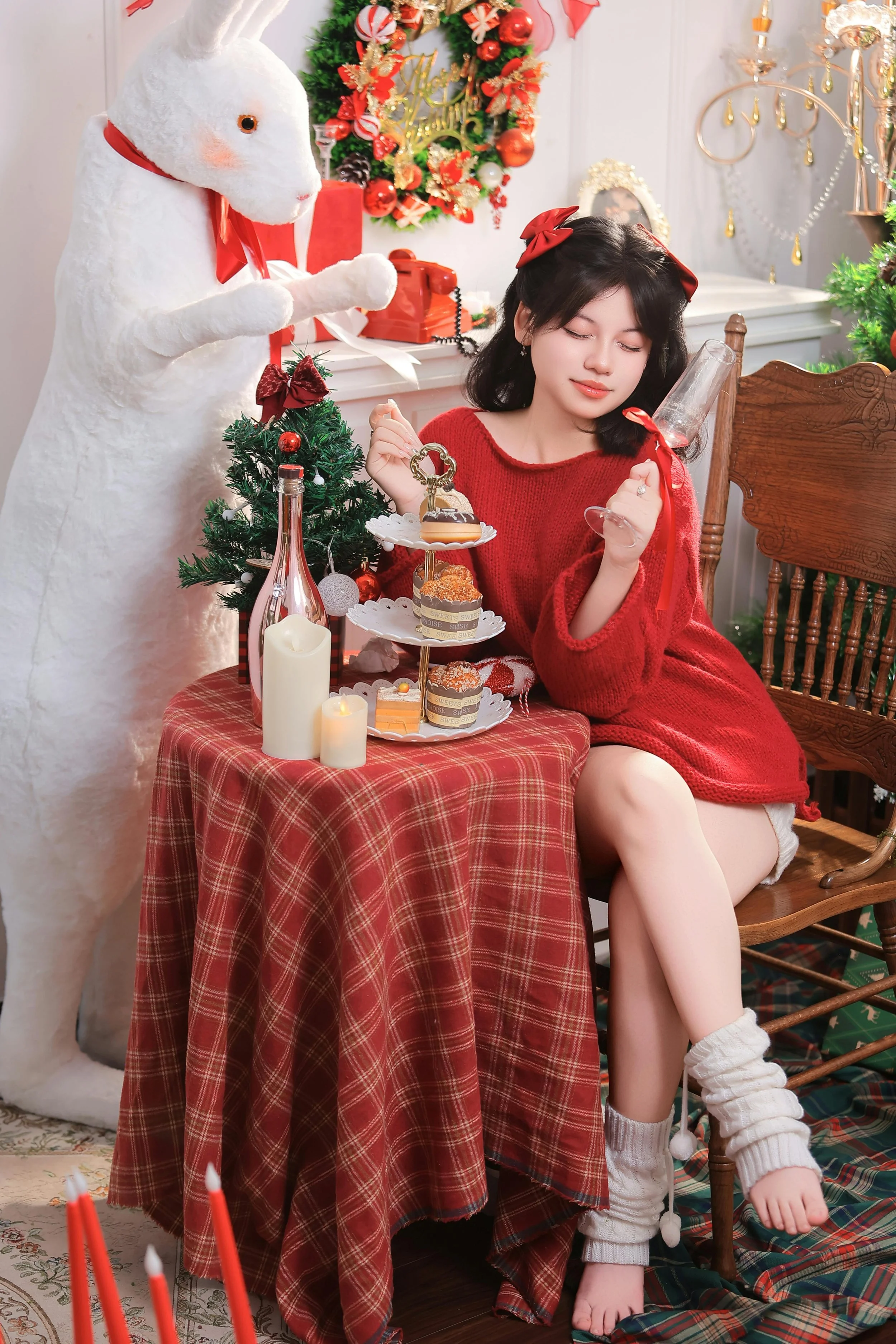 A young woman in a red sweater sitting at a table with a three-tiered dessert stand, candles, and holiday decorations, with a person in a bunny costume standing beside her in a festive Christmas setting.