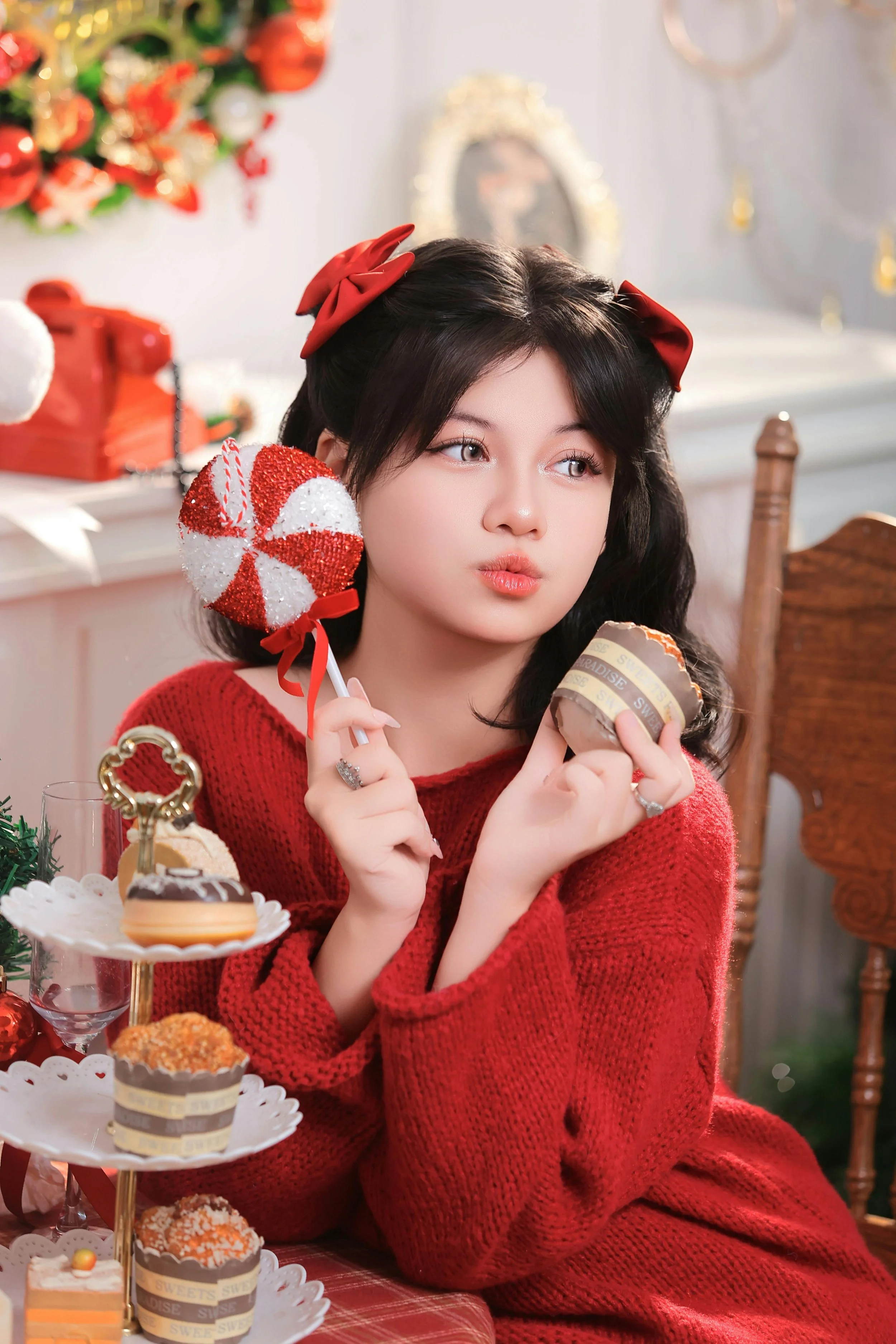 A young woman with dark hair wearing a red sweater and red bows in her hair, holding a peppermint candy and a cupcake, sitting at a decorated table with Christmas ornaments and treats.