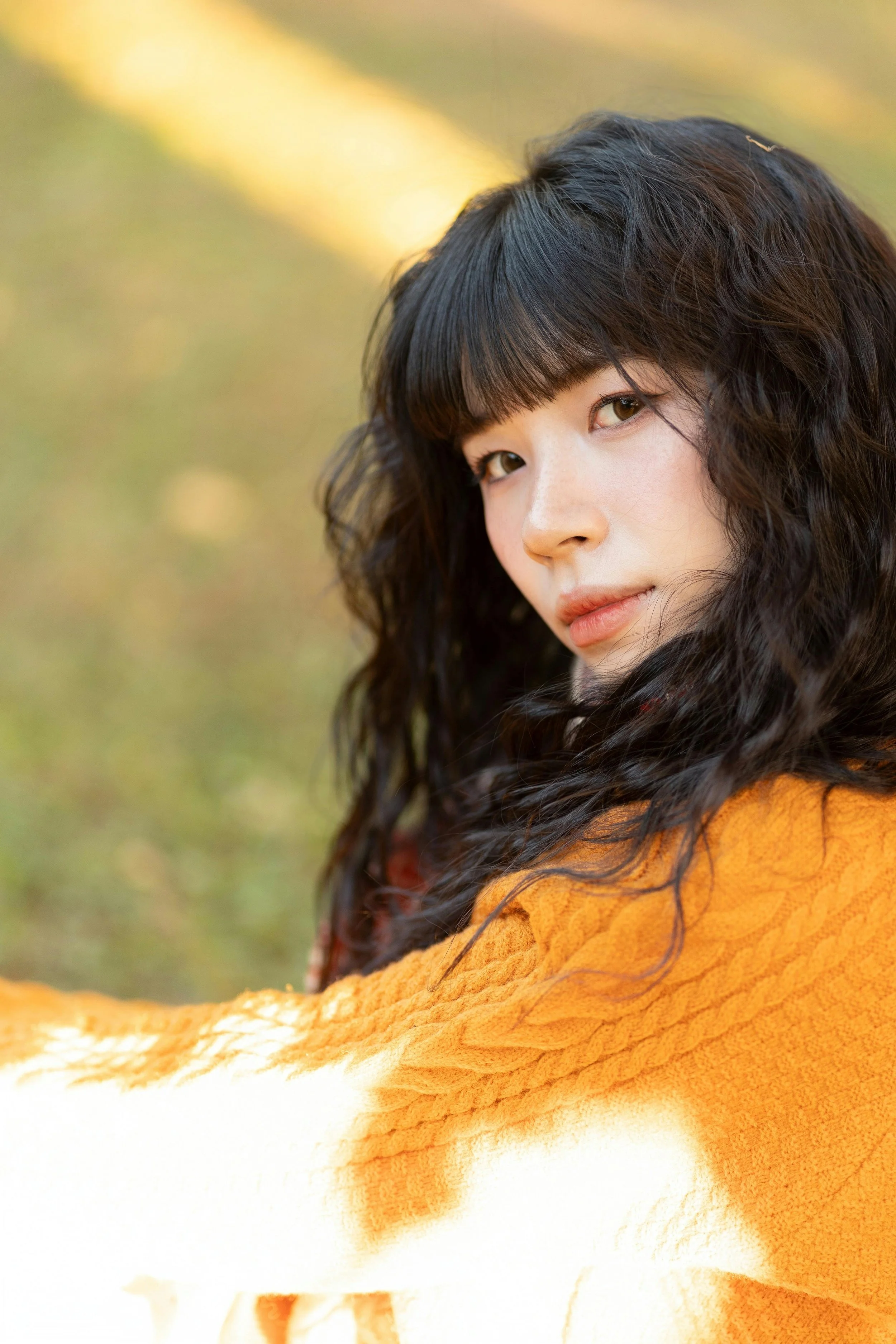 A young woman with long curly black hair and bangs looking at the camera outdoors with an orange and white blanket or sweater.