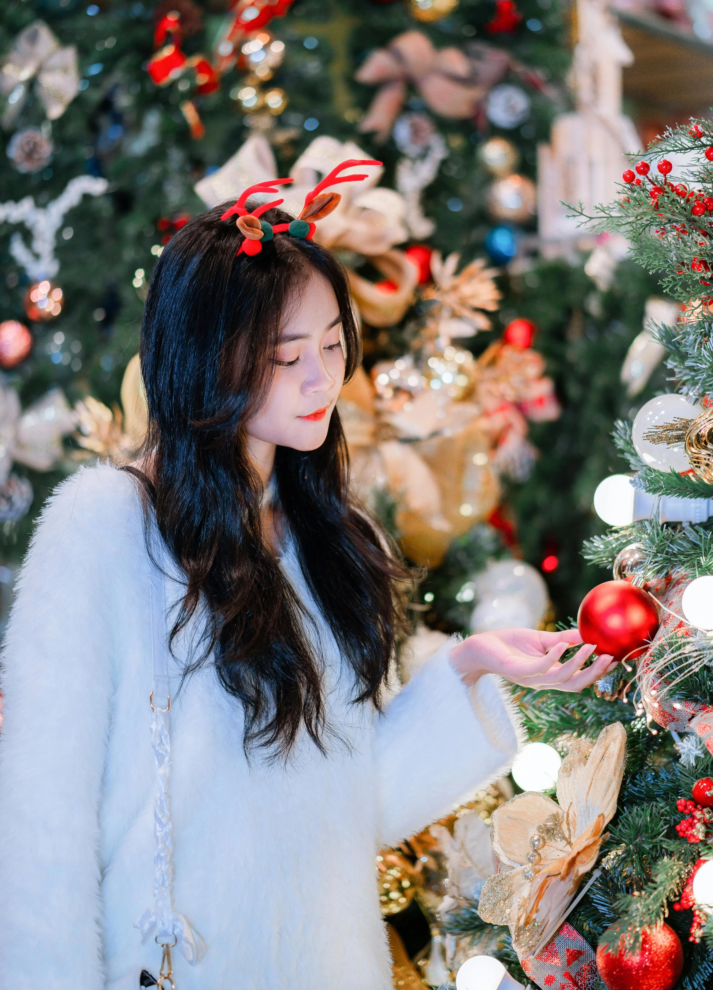 A woman with long dark hair dressed in a white fuzzy sweater with reindeer antler headband decorating a Christmas tree with ornaments.