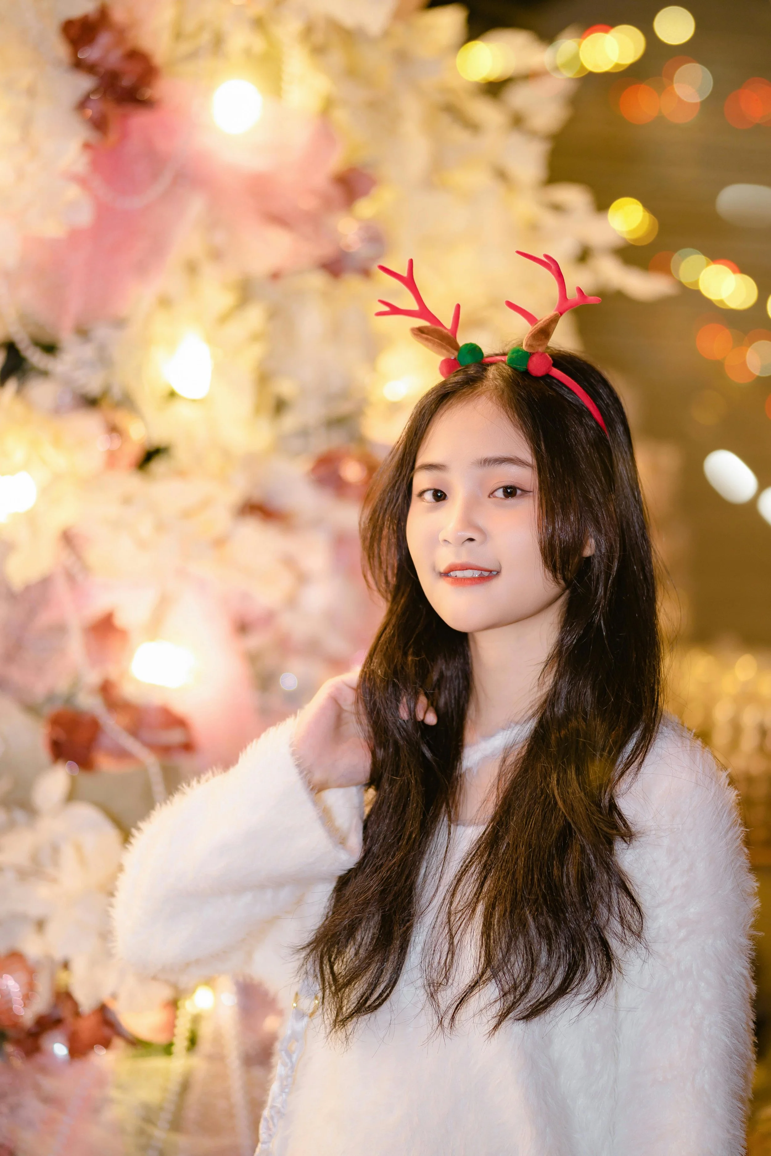 Young woman with long dark hair wearing reindeer antlers headband in front of decorated Christmas tree.