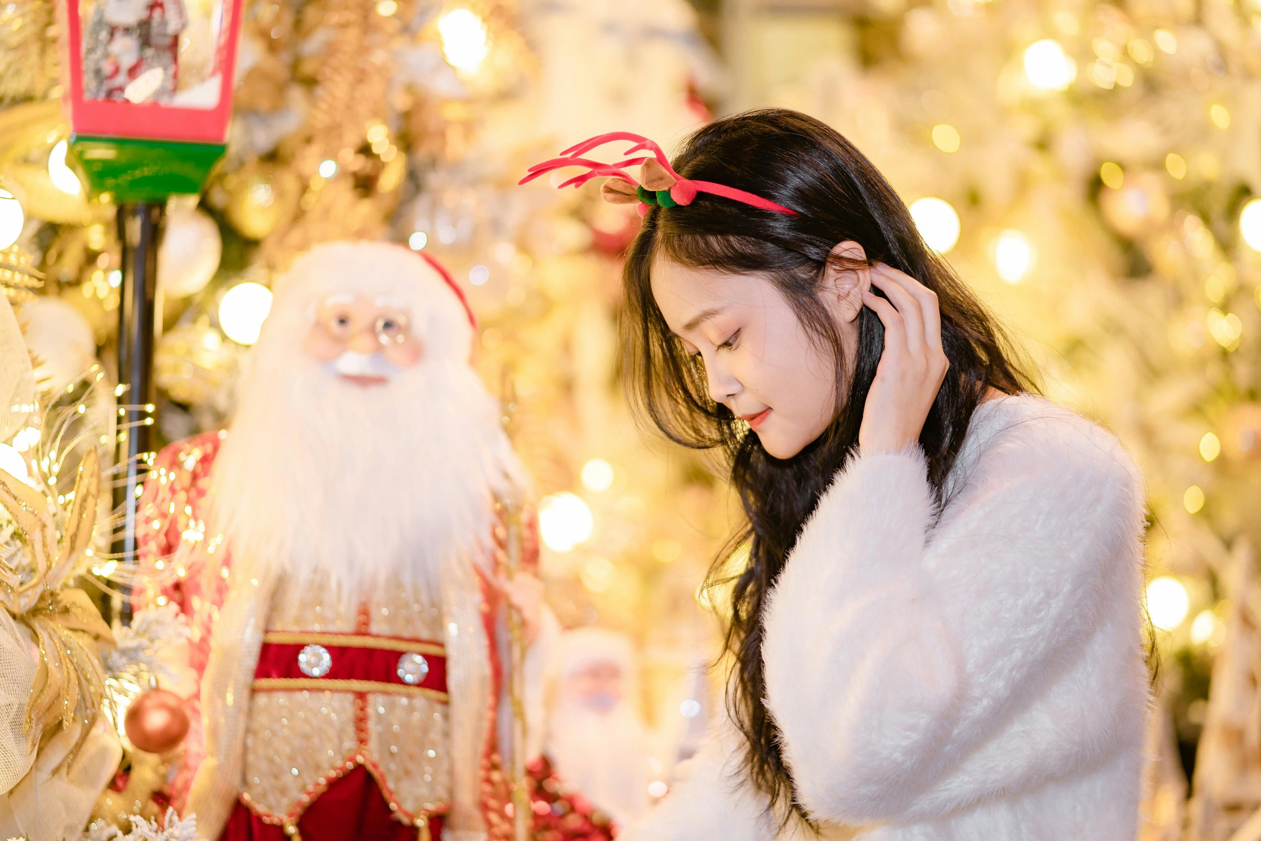 A woman dressed in a white fuzzy sweater, wearing a red Christmas headband with reindeer antlers, smiling with her eyes closed, is standing near a colorful Santa decoration surrounded by Christmas ornaments and lights.