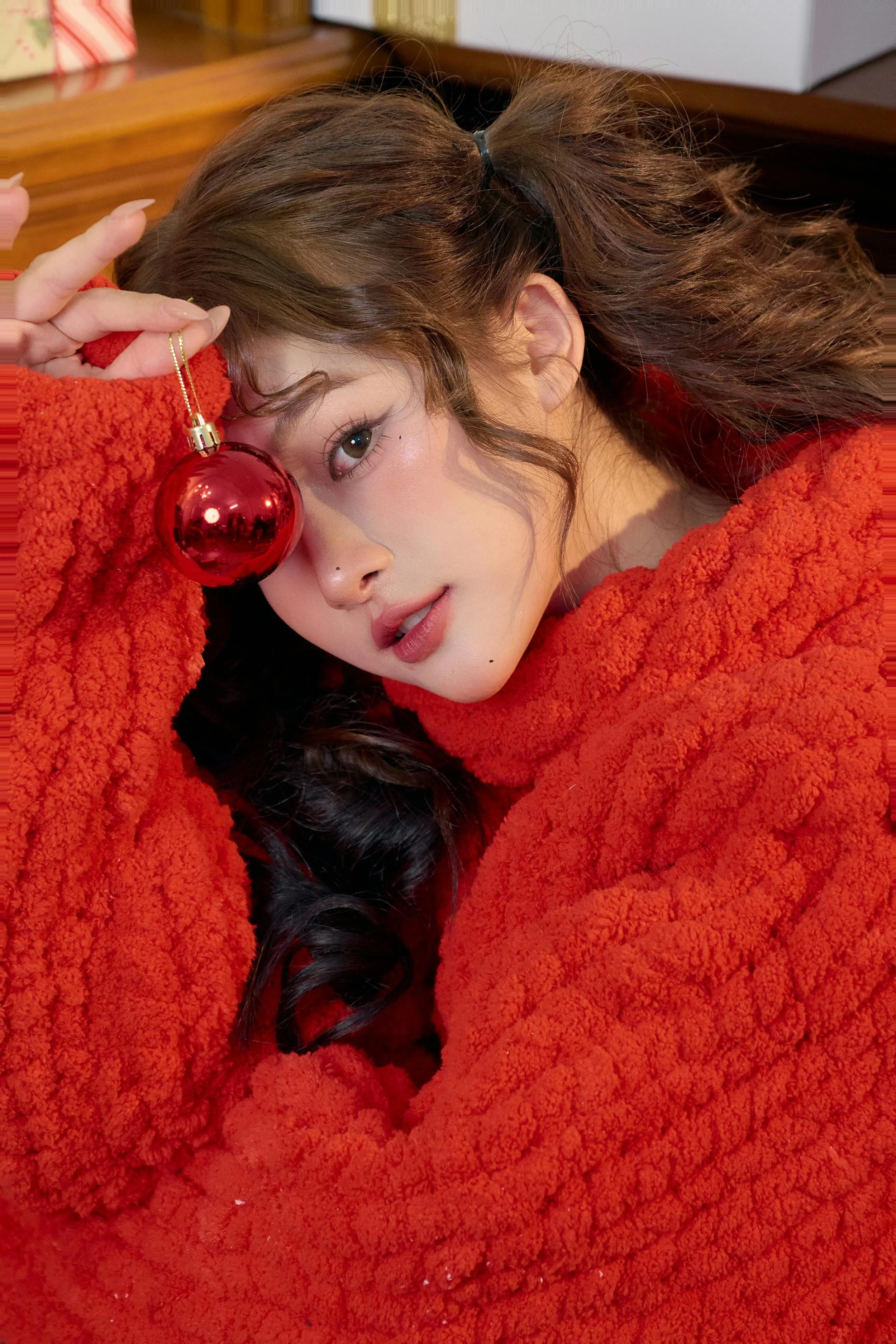 A woman with wavy brown hair in a red sweater holding a red Christmas ornament near her face, looking at the camera.
