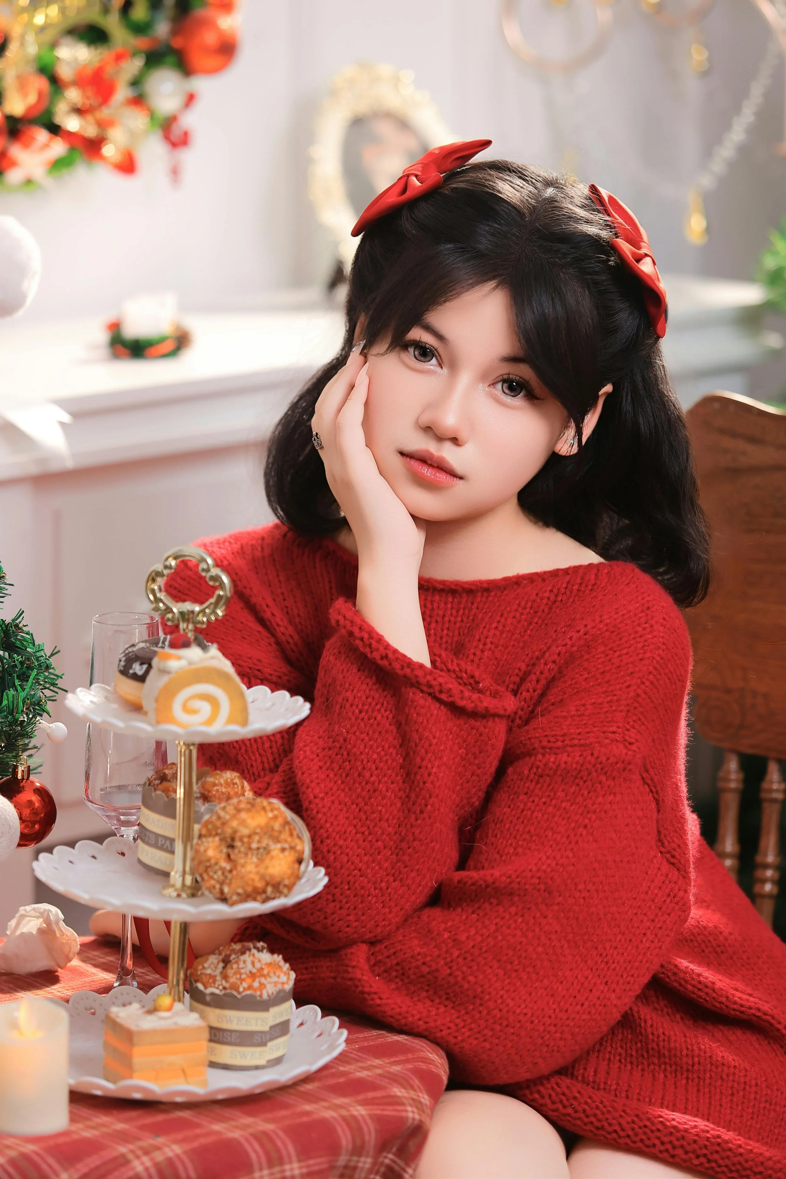 A young woman with black hair and a red bow headband wearing a red sweater, sitting at a table decorated for Christmas with a tiered dessert stand holding cookies and sweets, with Christmas decorations in the background.
