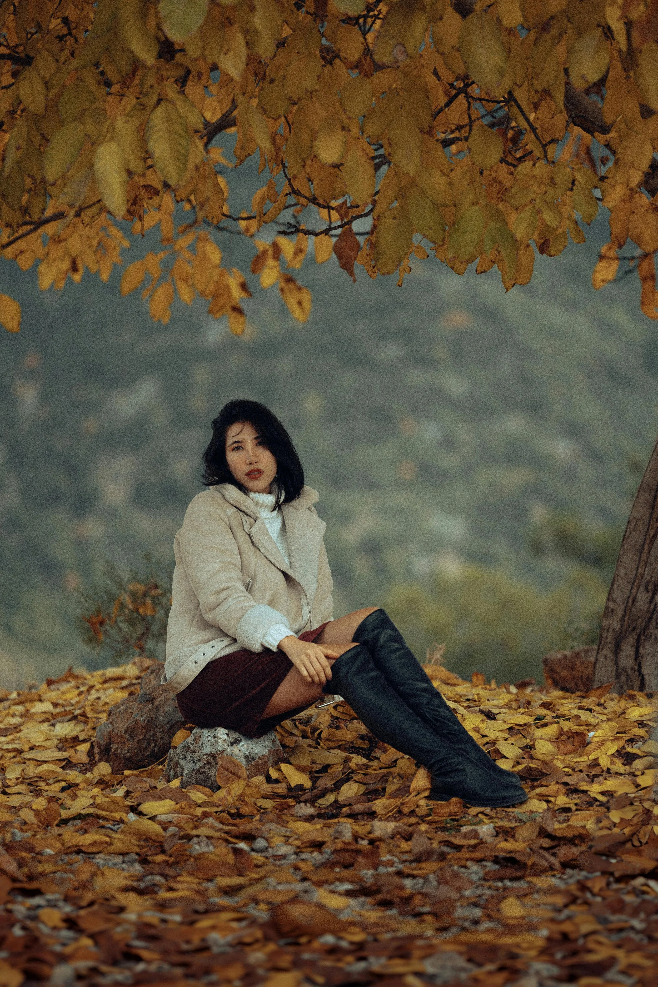 A woman sitting on a rock among fallen autumn leaves, wearing a beige coat and black over-the-knee boots, under a tree with yellow leaves.
