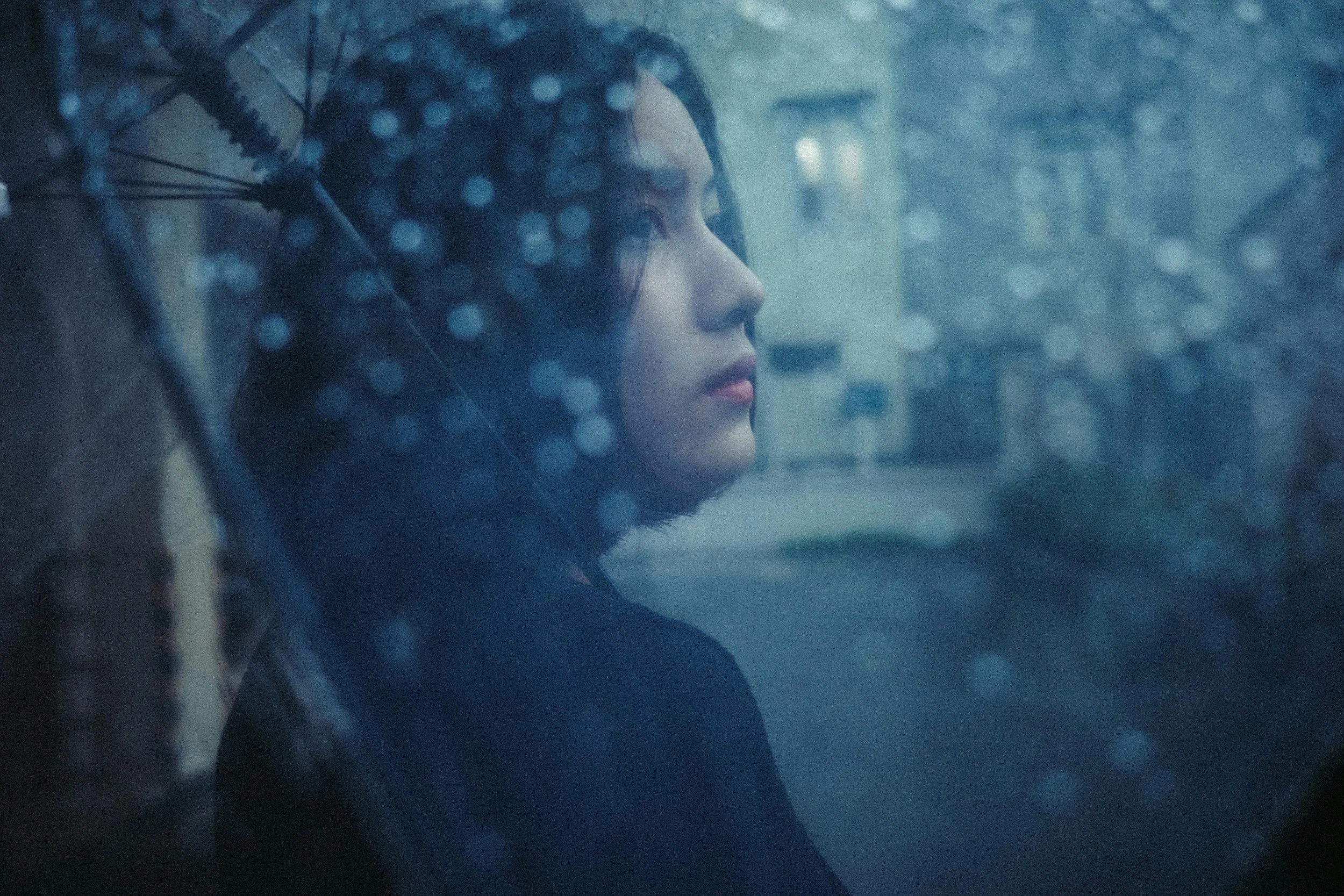 Woman with dark hair and fair skin sitting inside a car on a rainy day, looking out through a wet window.