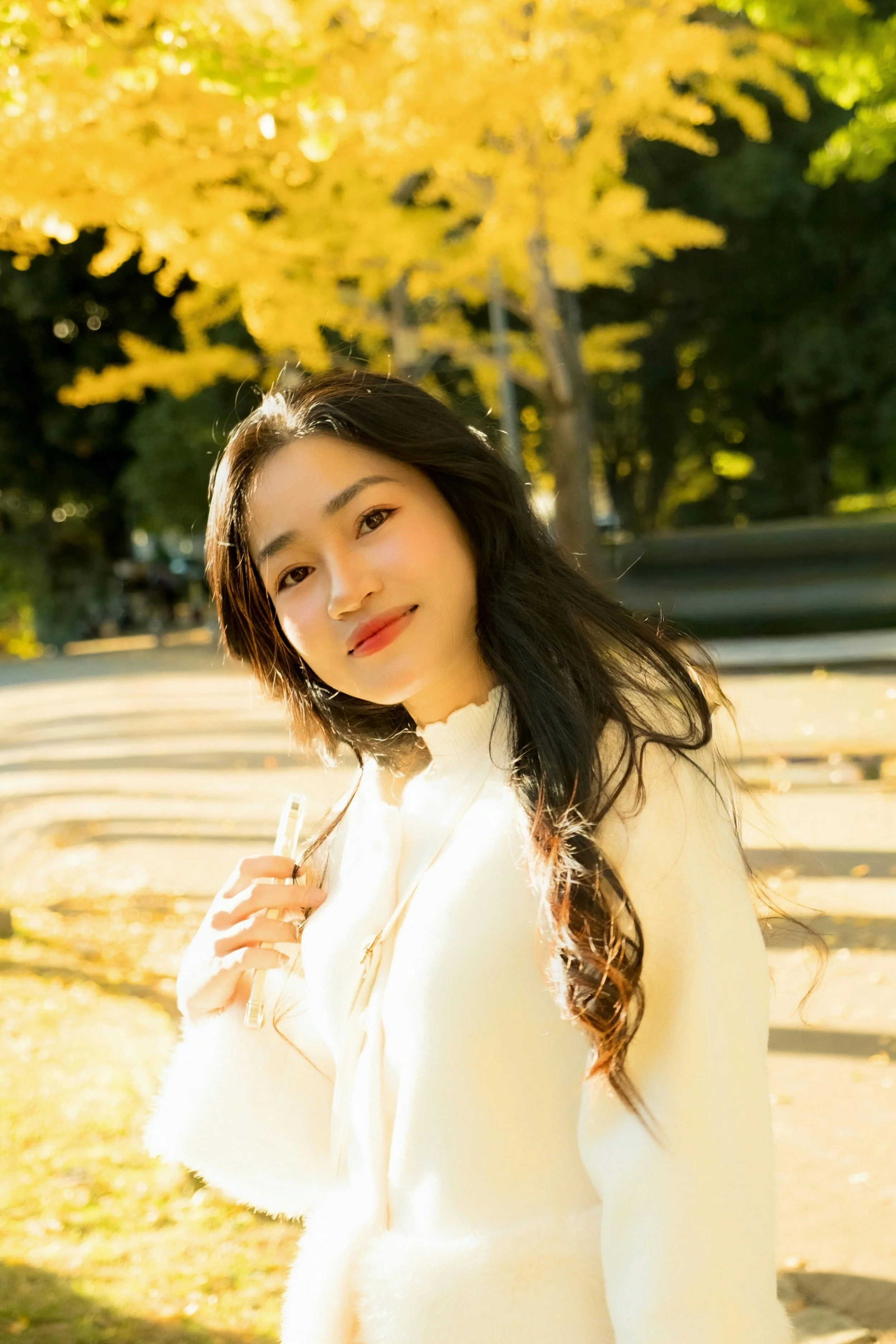 A young woman with long dark hair smiling outdoors in a park during fall, holding a pen, with yellow autumn leaves in the background.