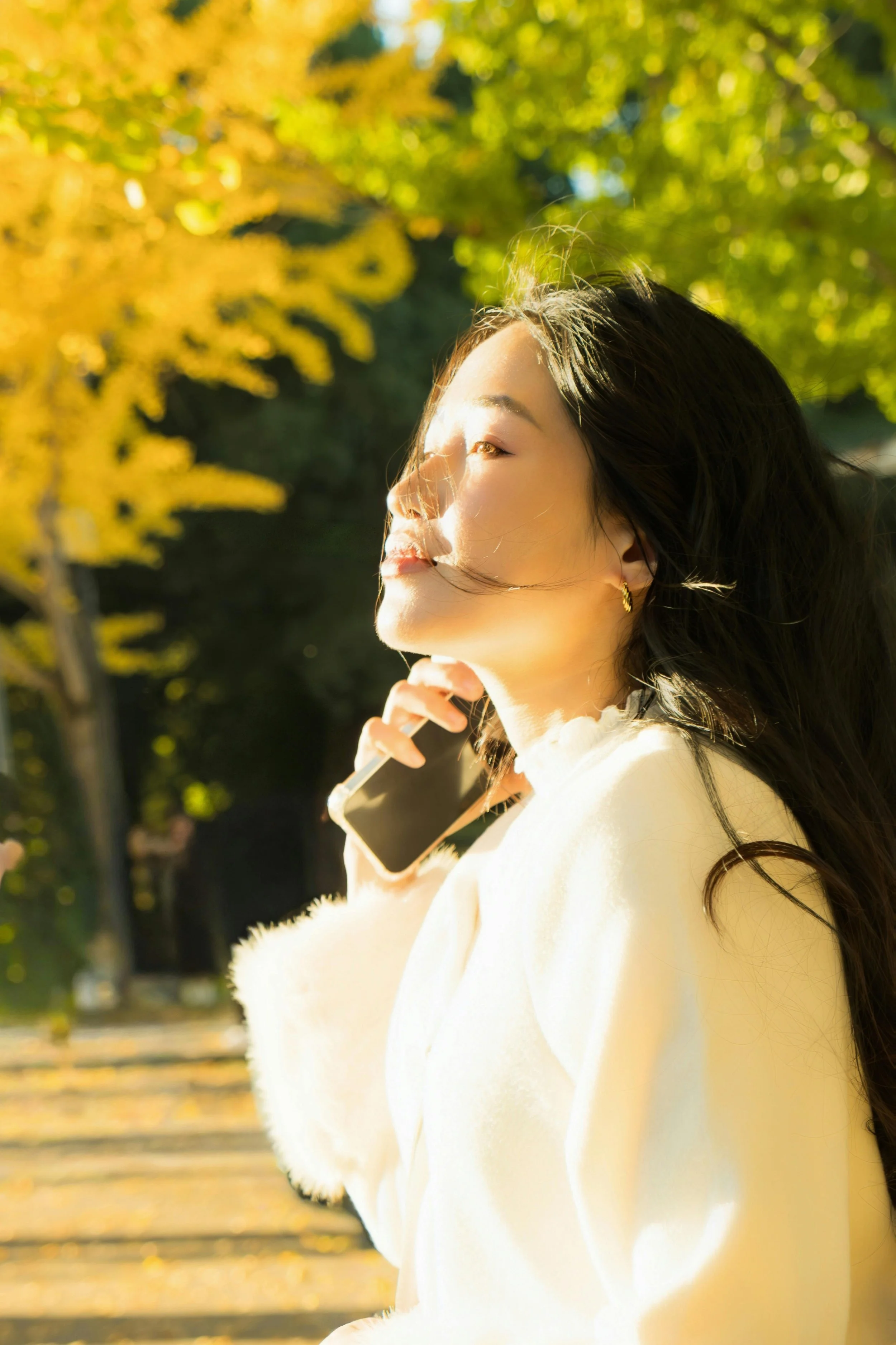 A woman outdoors talking on her cell phone in a park with yellow and green autumn trees.