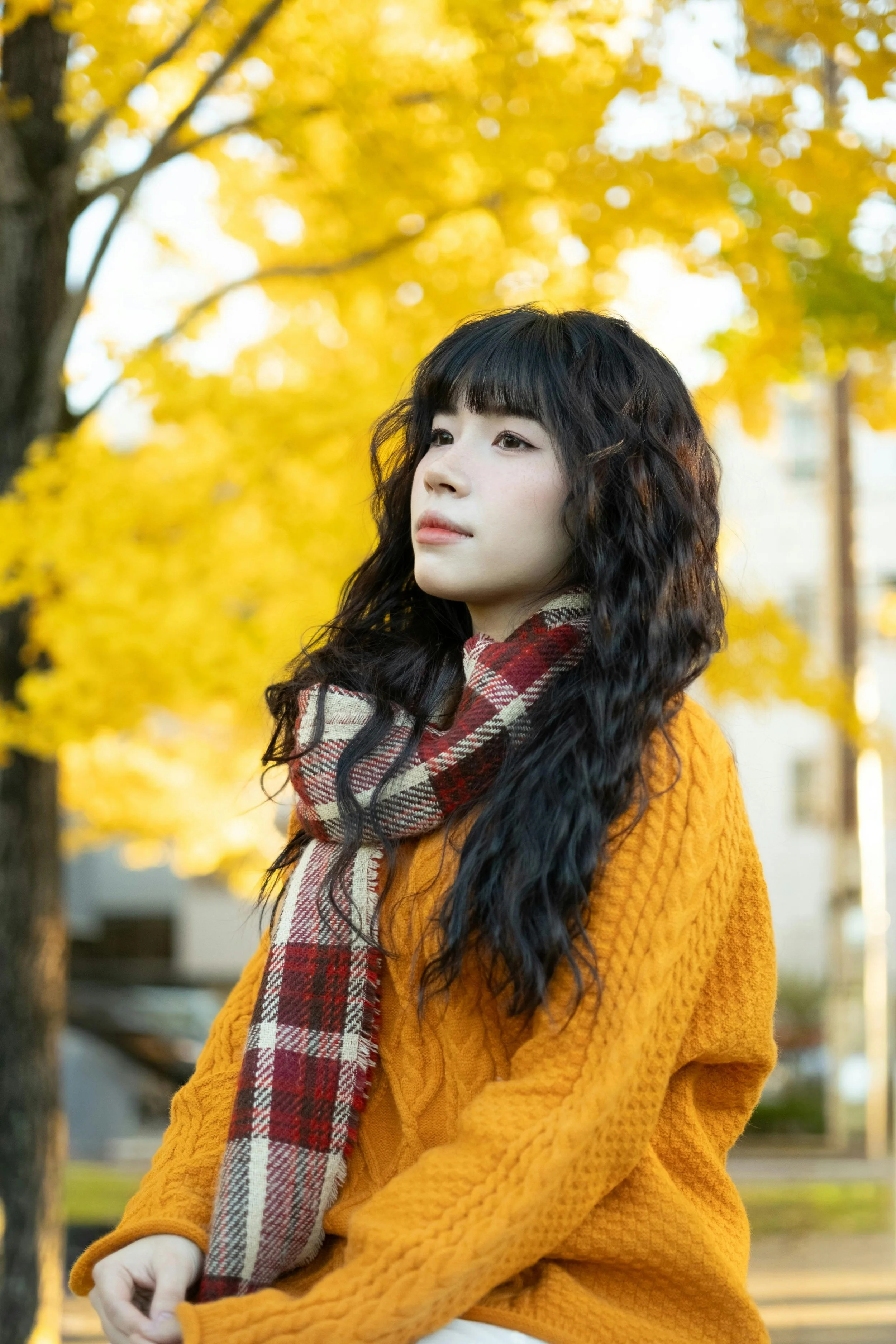 A woman with long curly black hair and bangs standing outdoors in front of yellow autumn trees, wearing a yellow sweater and a red plaid scarf.