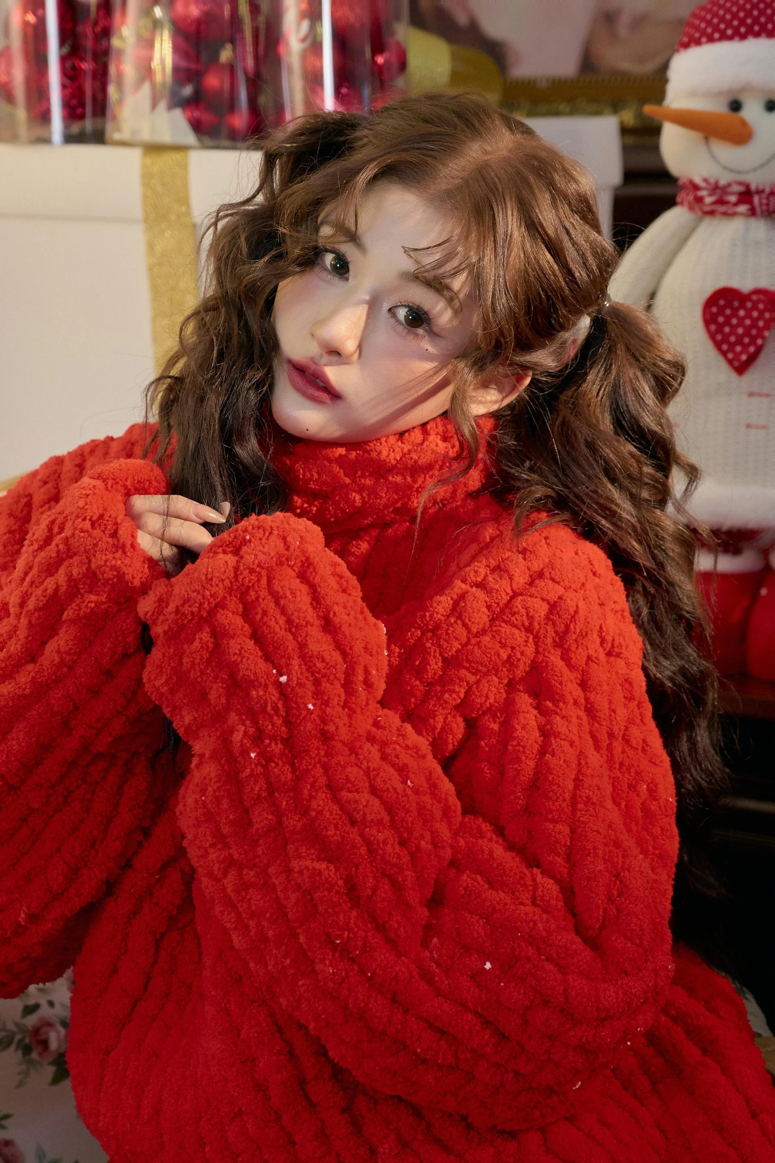 A woman with long, curly brown hair and fair skin wearing a bright orange, cozy, fleece sweater, posing indoors with a Christmas decoration background, including a snowman with an orange carrot nose, black eyes, a red scarf, and a polka dot heart on 