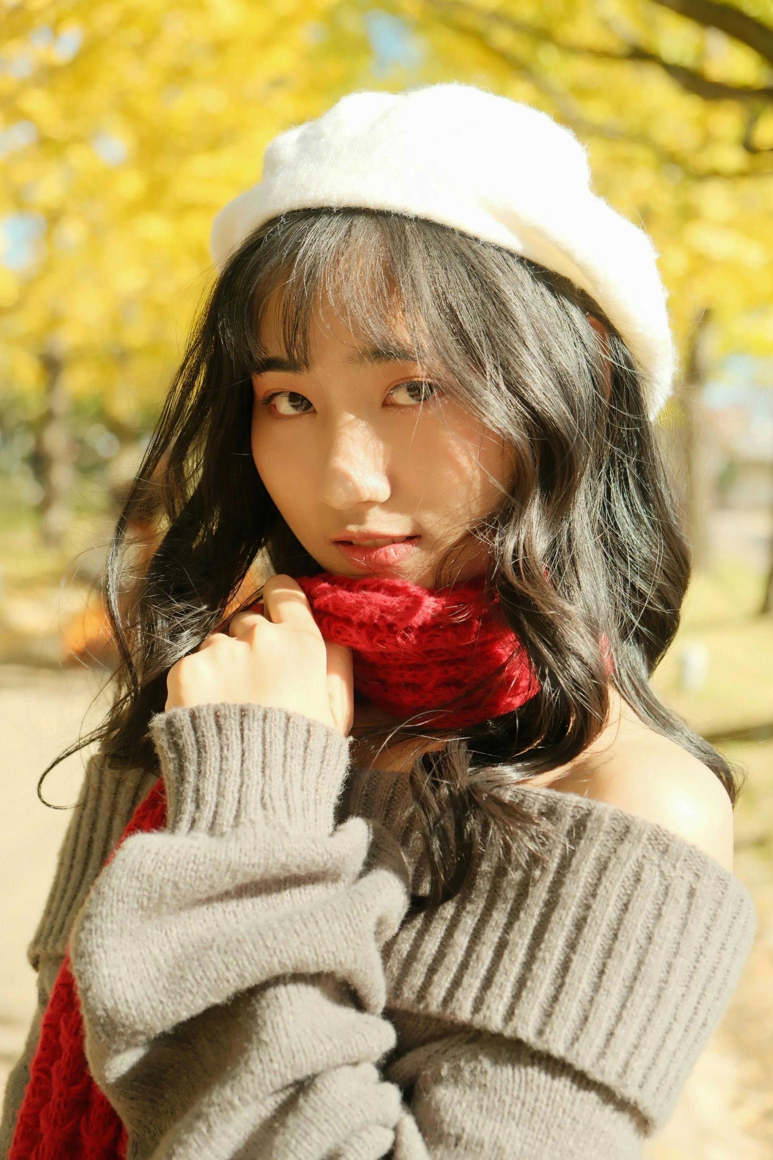A young woman with long dark hair wearing a cream sweater, red scarf, and white beret outdoors in a park during autumn with yellow leaves.