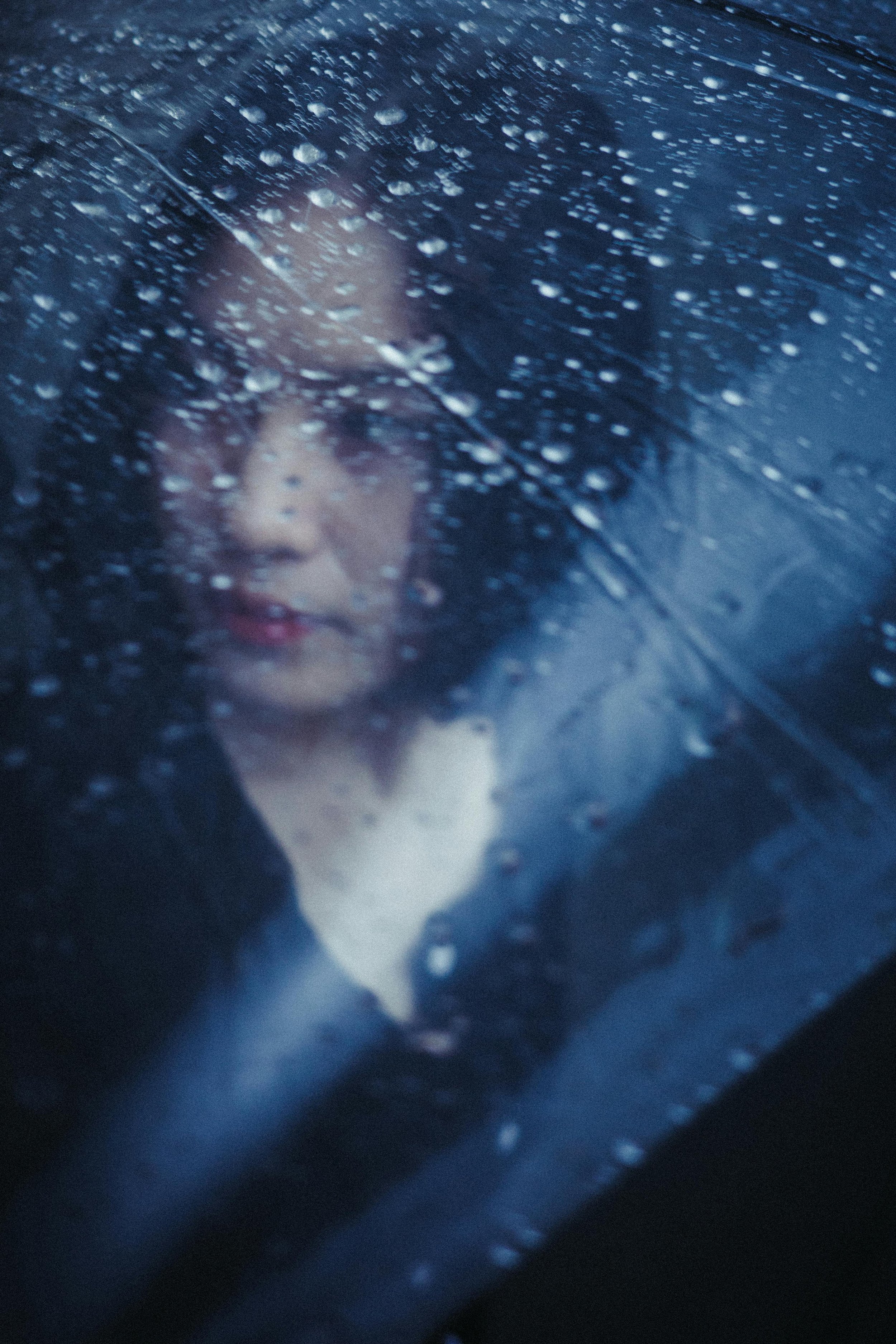 A woman with dark hair looking out through a rain-covered window on a dark day.