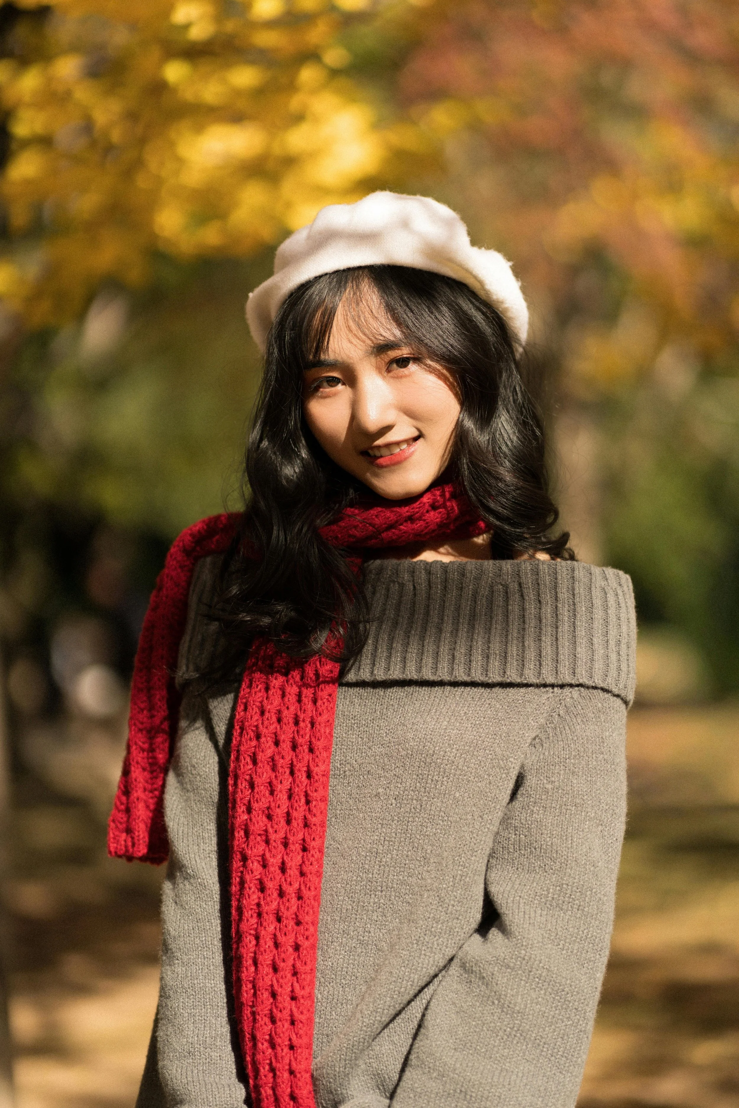 A woman smiling in an outdoor setting during autumn, wearing a white beret, a red scarf, and a gray sweater.