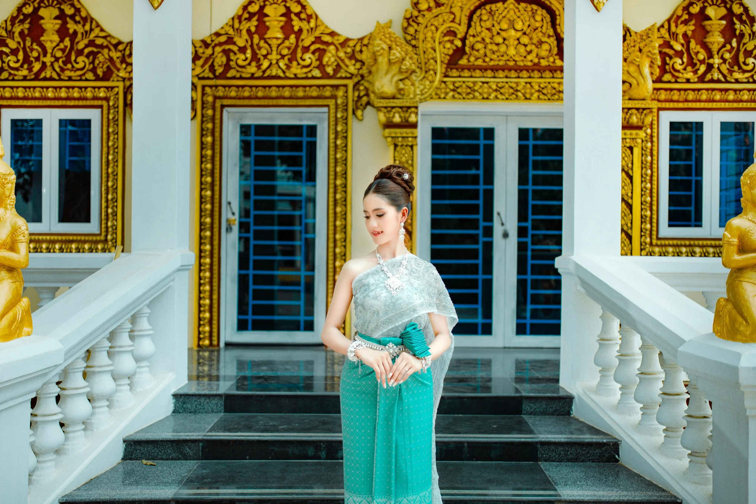 A woman dressed in traditional Thai attire, standing on the steps of an ornately decorated building with gold accents and blue-tinted windows.