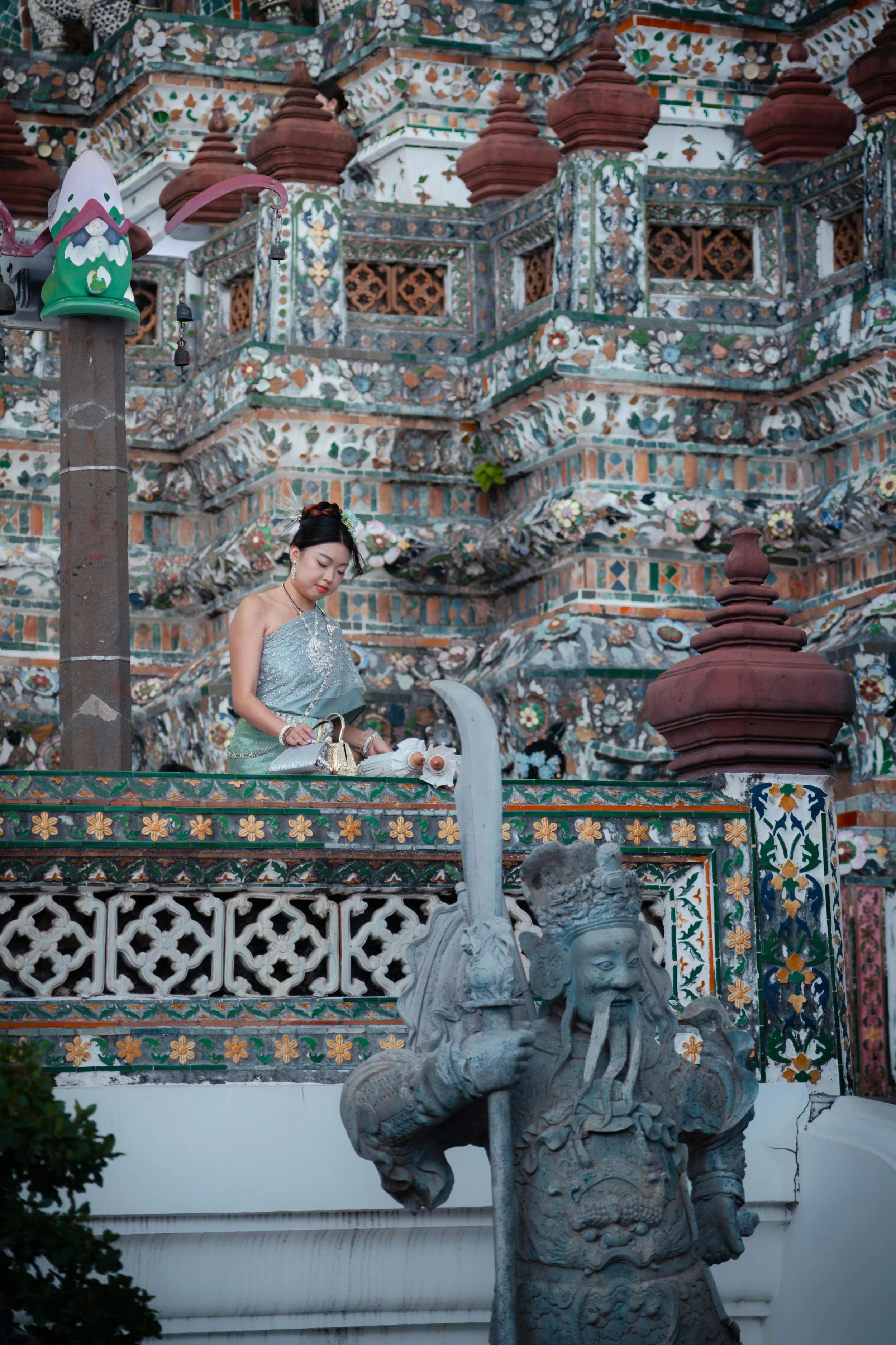 A woman in traditional attire standing on a decorated platform at a Buddhist temple, with elaborate floral mosaic walls and a stone guardian statue in the foreground.