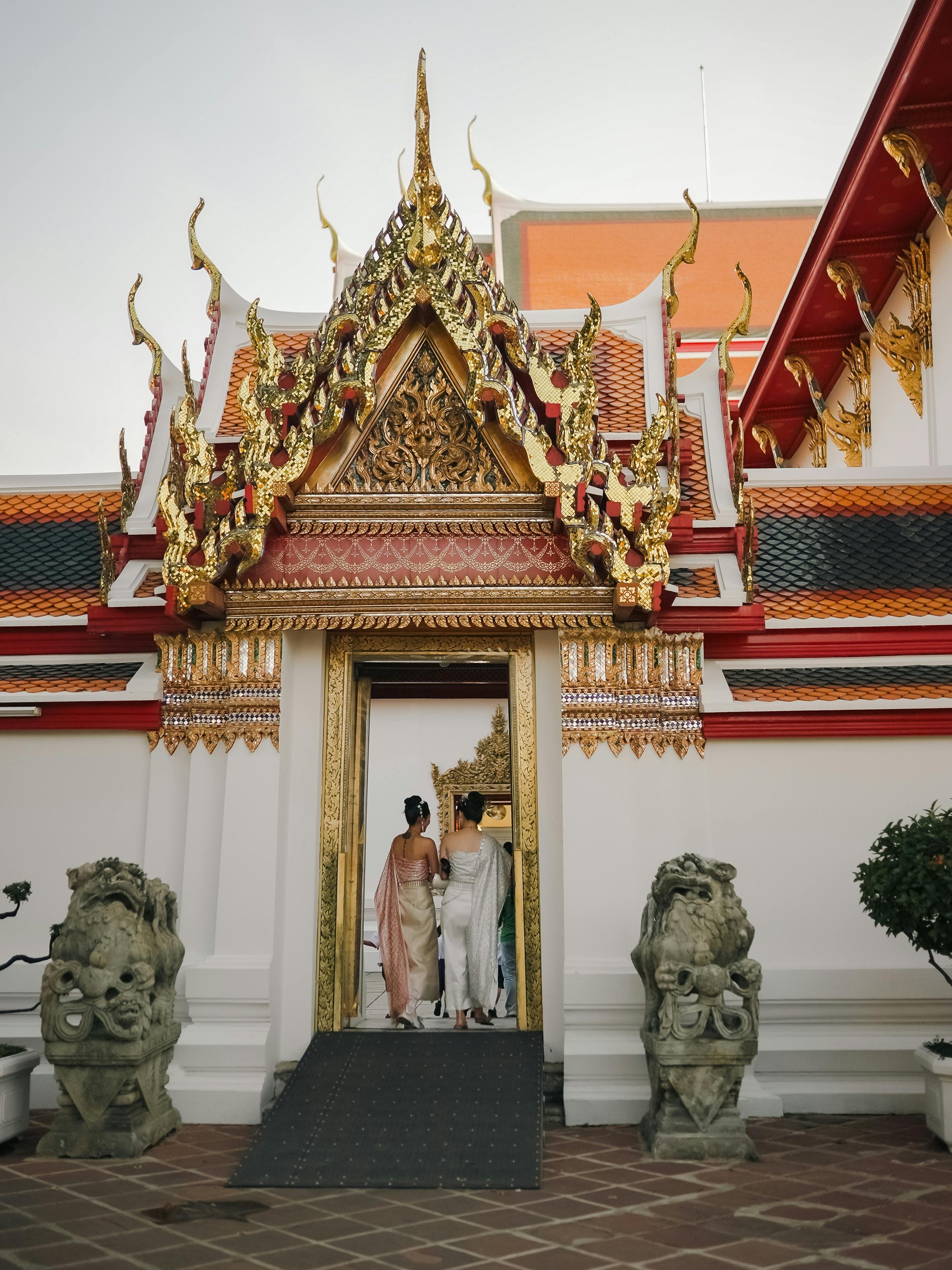 Two women dressed in traditional Thai attire walking into a Thai temple with ornate gold decorations and statues.