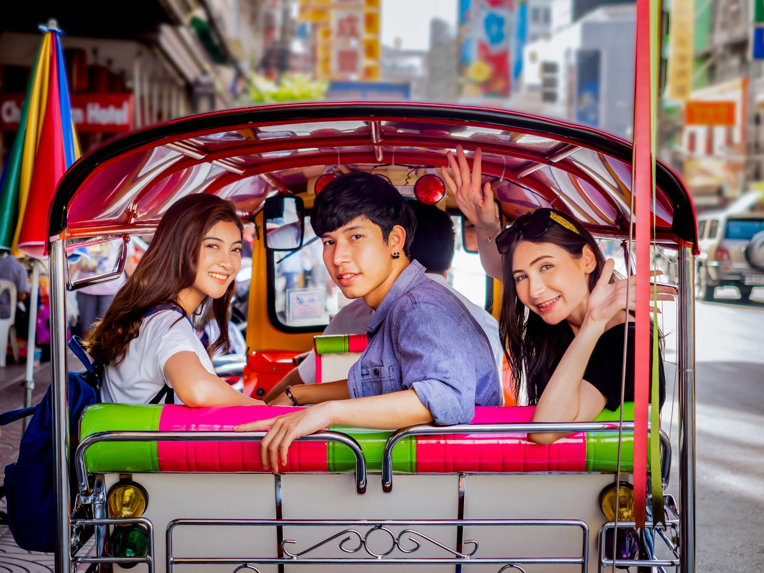 Three young people sitting in a colorful tuk-tuk on a busy city street with vibrant signs and vehicles in the background, smiling and waving.