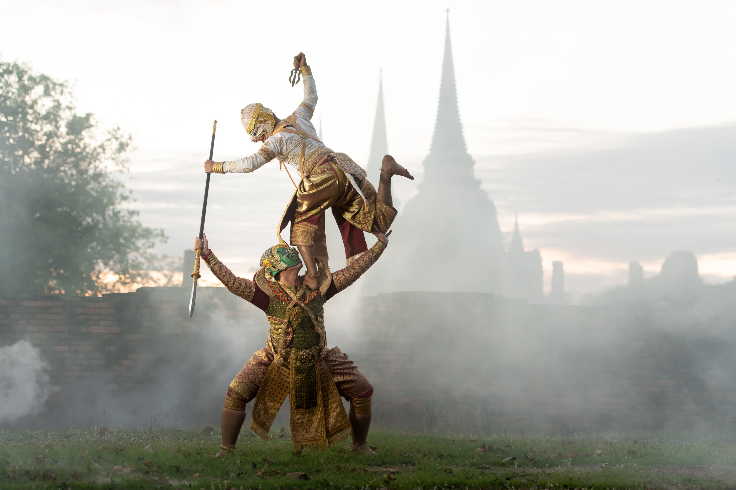 Traditional Thai puppetry performance featuring three performers in ornate costumes and masks, with a temple and stupas in the background at sunrise or sunset.