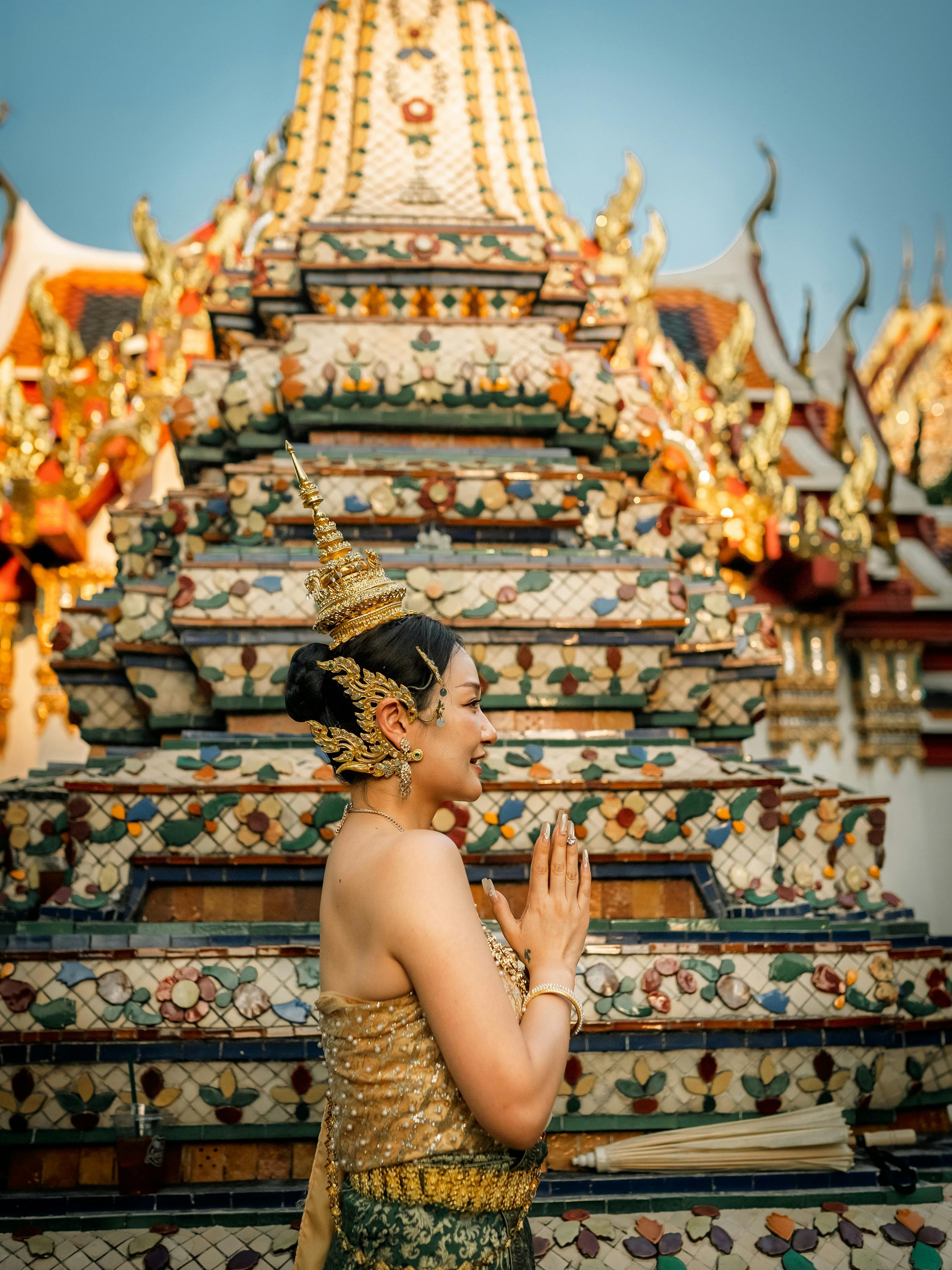 A woman in traditional Thai attire praying in front of an ornate temple decorated with colorful mosaic tiles and gold accents.