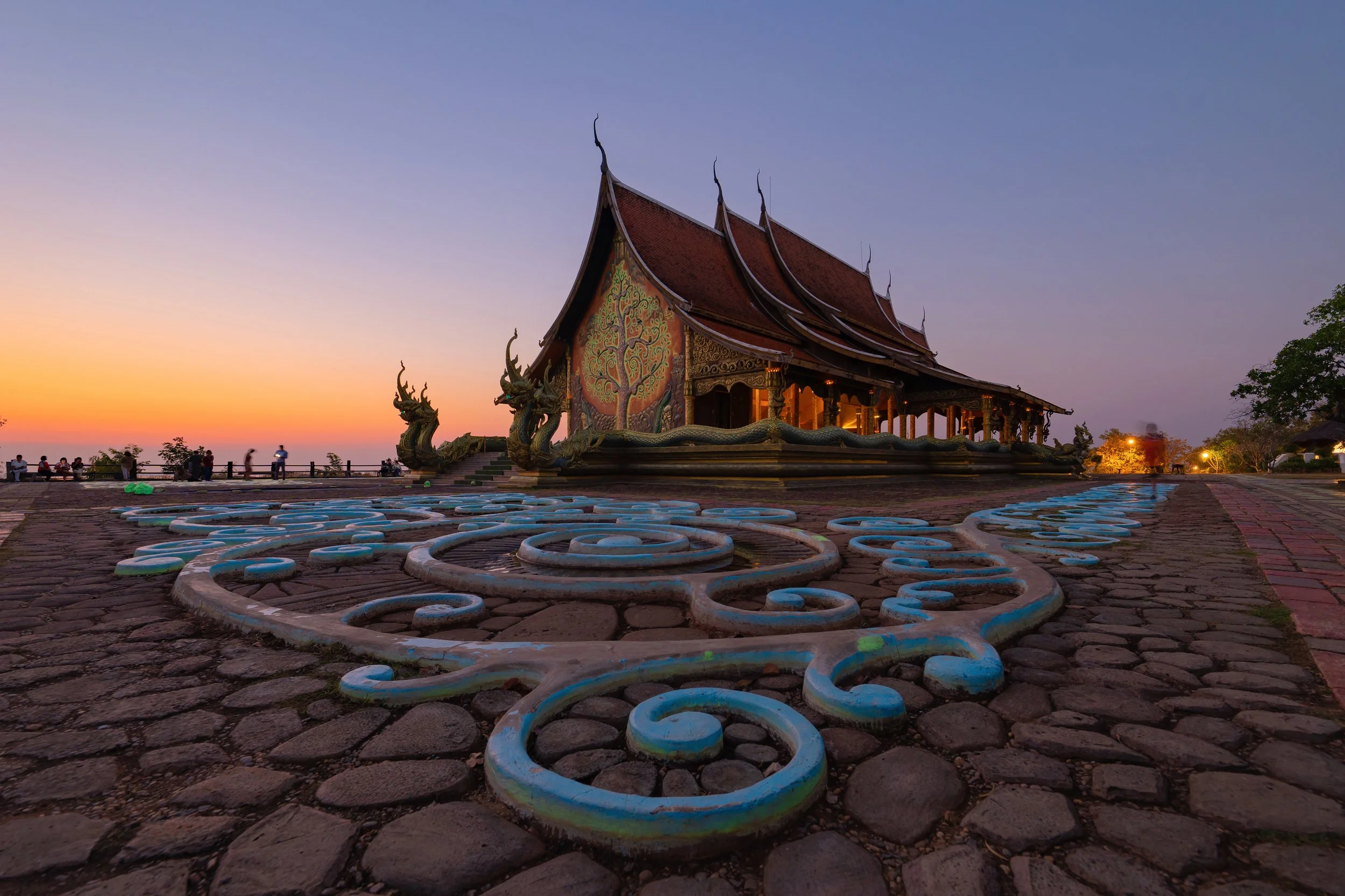 Traditional Thai temple with ornate decoration, dragons at the entrance, and a brightly colored mural on the wall, during sunset with people in the distance.