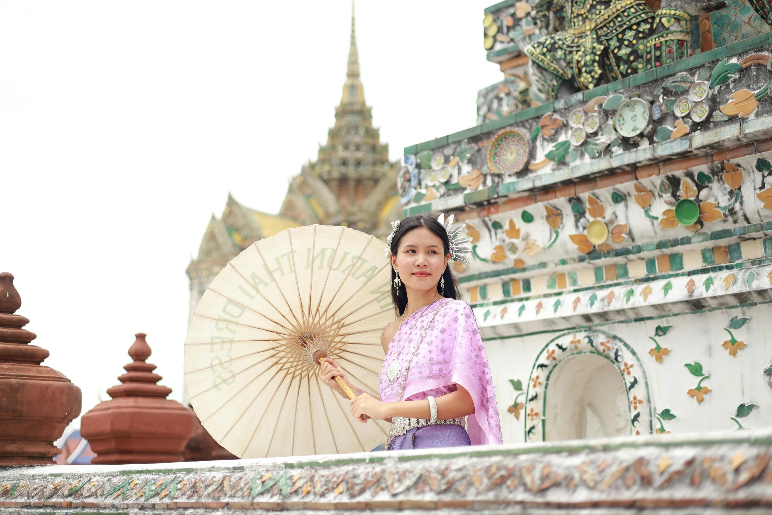 A young woman dressed in traditional Thai attire holding a parasol, standing in front of an ornate and colorful temple wall decorated with floral and bird motifs.