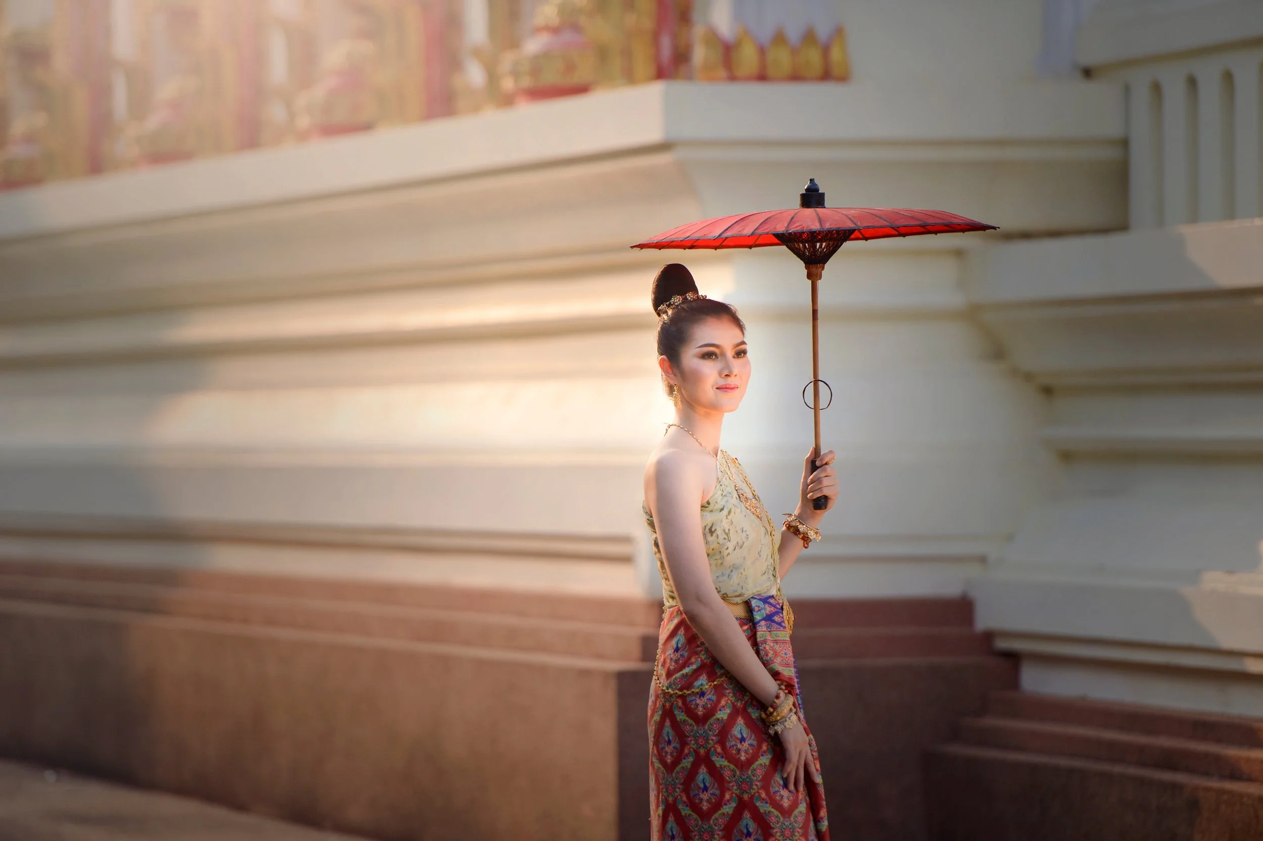 A woman dressed in traditional Thai attire holding a red parasol standing near a white structure, with warm sunlight creating a soft glow.